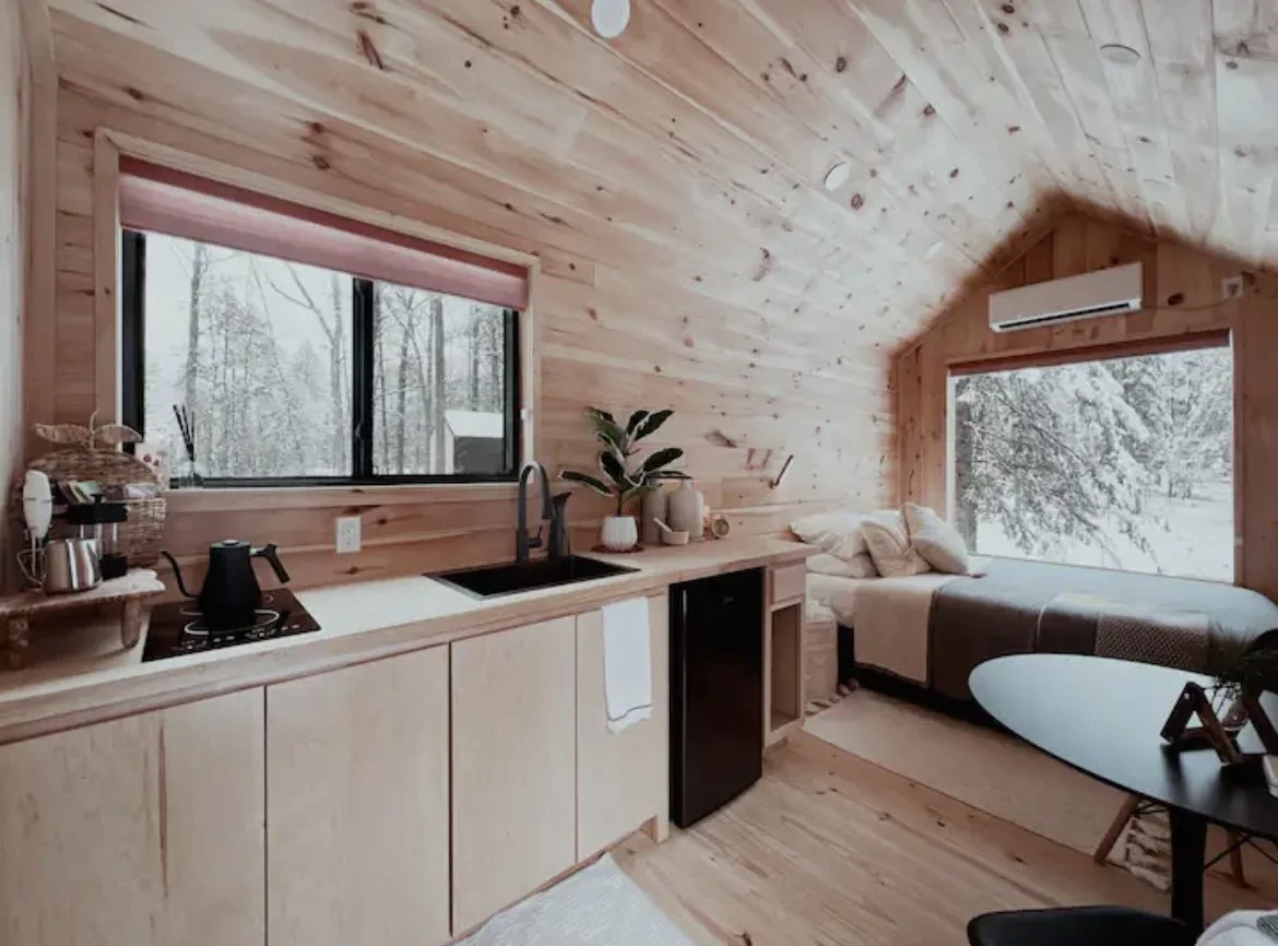 A cozy living space with wood-paneled walls and ceiling. There is a small kitchen area with a window showing snow-covered trees outside. The kitchen has a white countertop, a black sink, a stove, and a black mini fridge. Next to the kitchen is a daytime bed with pillows, positioned beneath a large window showing snow-covered trees. A potted plant, decorative items, and a white towel hang from the cabinet. A small black table with a surfboard leaning against it is also visible.