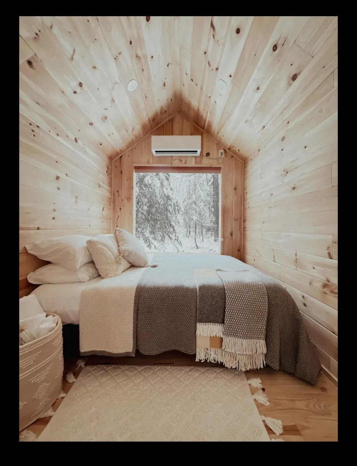 A cozy bedroom with wood-paneled walls and ceiling. There is a bed with white pillows, beige and brown blankets, and a basket of towels nearby. A large window behind the bed shows a snowy outdoor scene with trees.