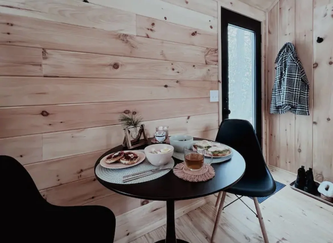 A cozy dining nook with a round black table set for breakfast, featuring bowls, plates with bagels or toast, and a cup of tea, against a wood-paneled wall with a coat hanging on the wall near a glass door.