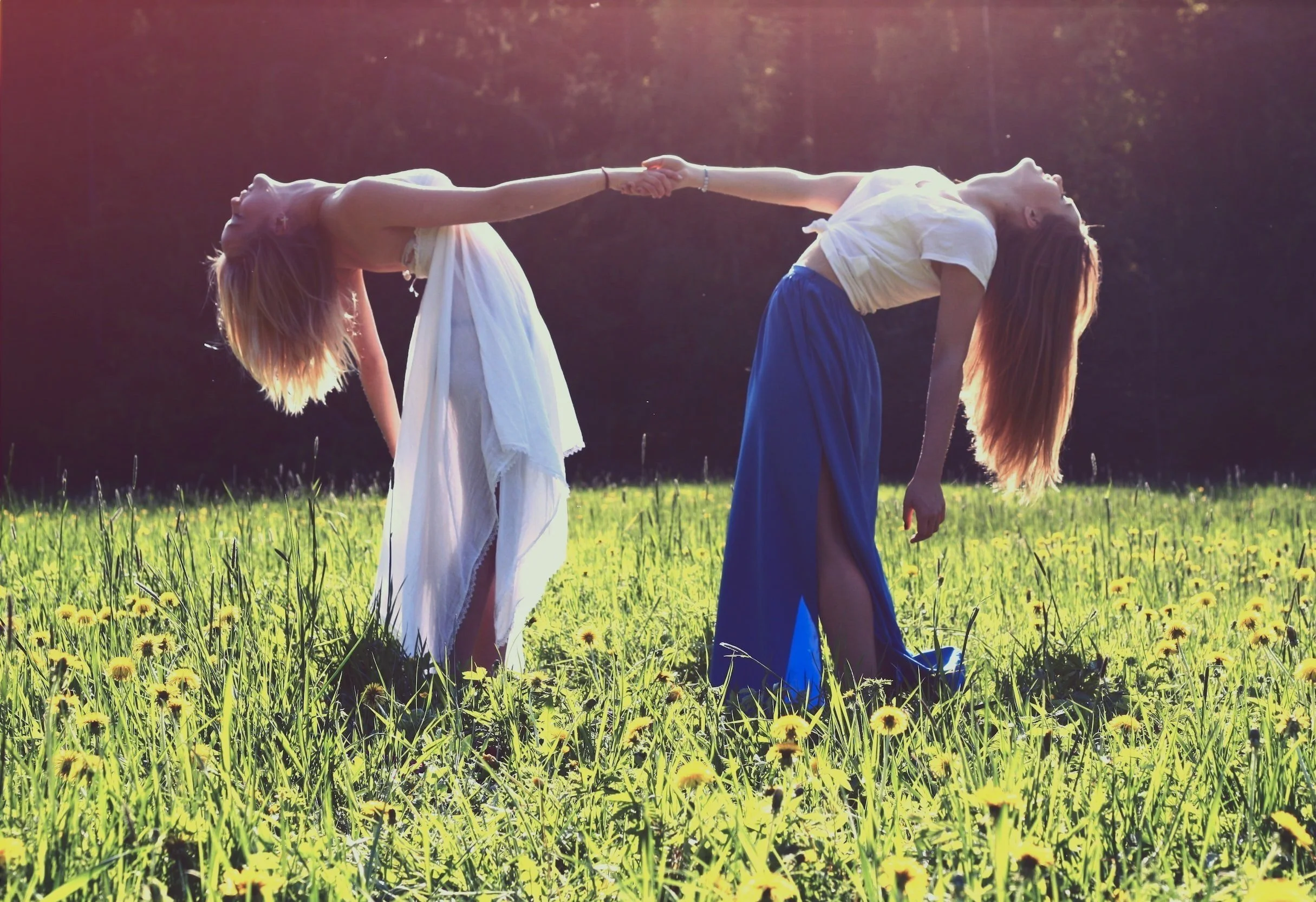 Two women with long hair doing a yoga pose in a grassy field during sunset, holding hands and bending backward.