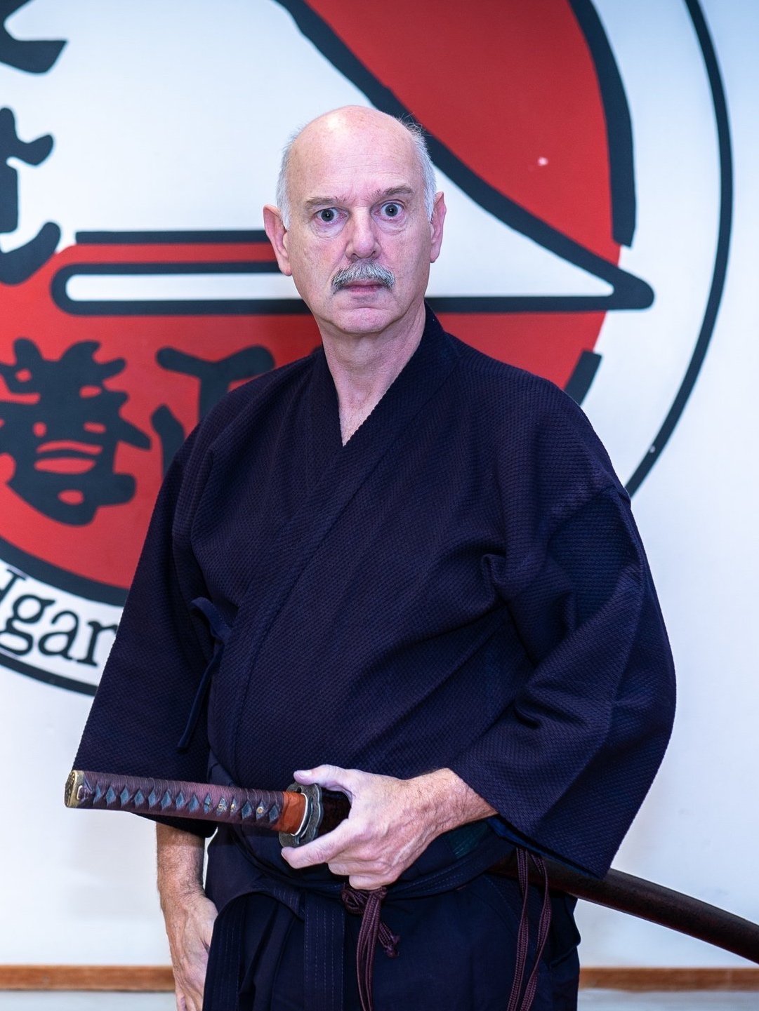 Een man in een zwarte martial arts-gewas houdt een katana vast voor een bord met een Japans symbool.