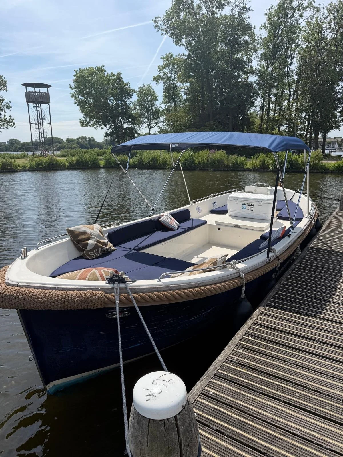 Een motorboot ligt aan de kade van een waterweg. De boot heeft een blauwe kap en is uitgerust met kussens en een koelbox. Op de achtergrond staan bomen en een uitzicht over het water met een toren in de verte.