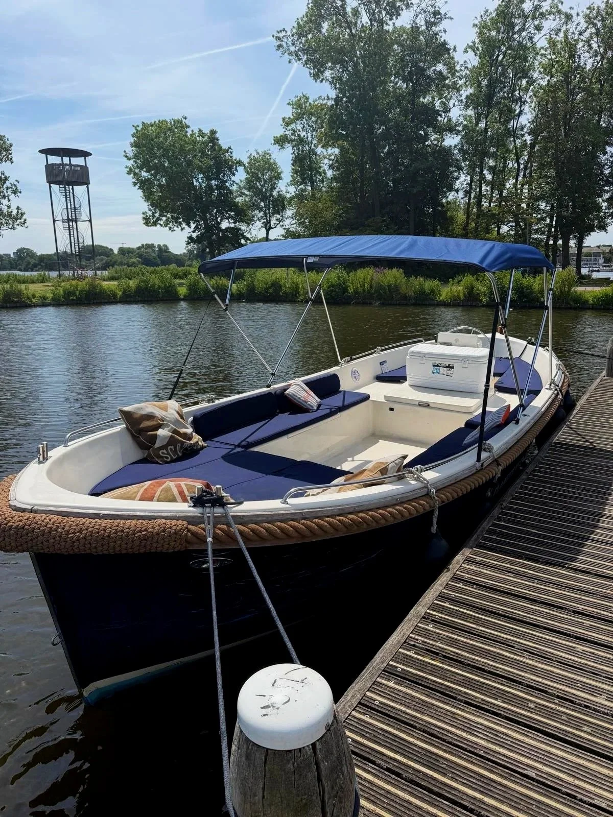 Een wit motorboot met een blauwe zonnedakje ligt aan een houten steiger langs een rivier, met bomen en een uitkijktoren op de achtergrond.