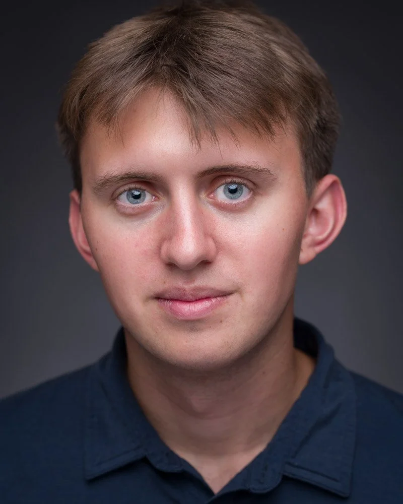 Actor headshot of a young man with light brown hair and blue eyes wearing a dark blue collared shirt, looking directly at the camera with a neutral expression.