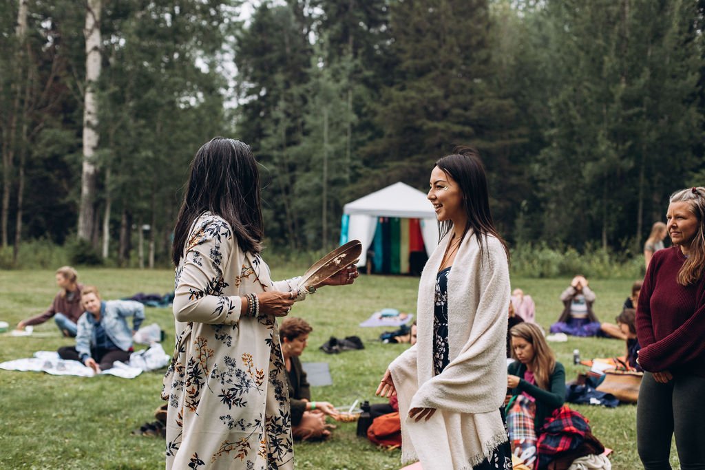 Outdoor gathering with women conversing, surrounded by people sitting on blankets, with a tent and forest in the background.