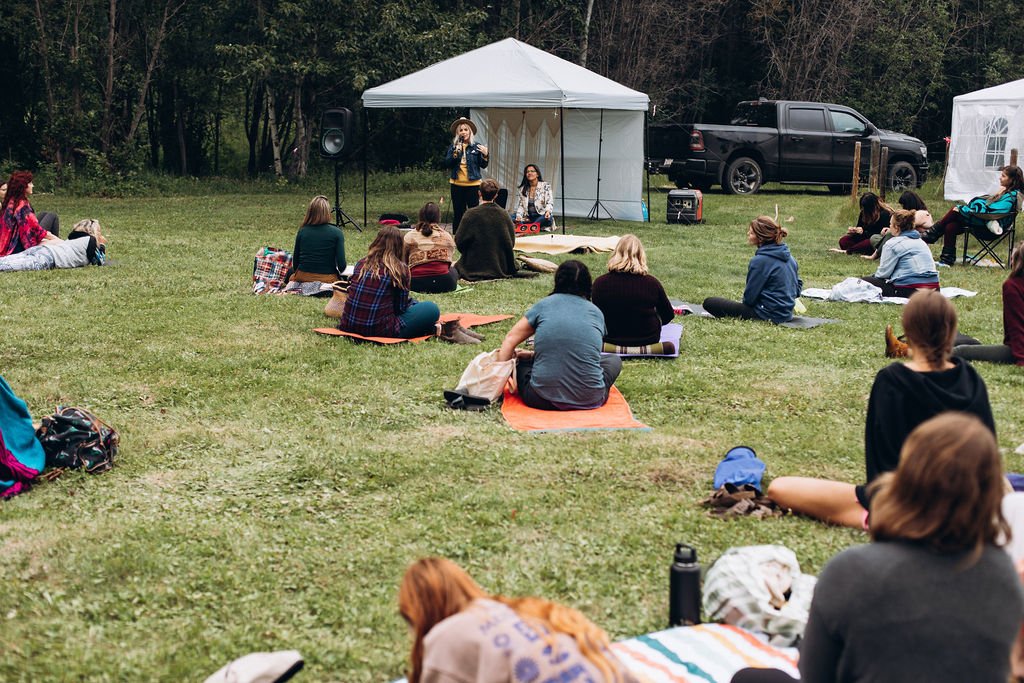 Outdoor event with people sitting on grass listening to a speaker under a canopy tent.
