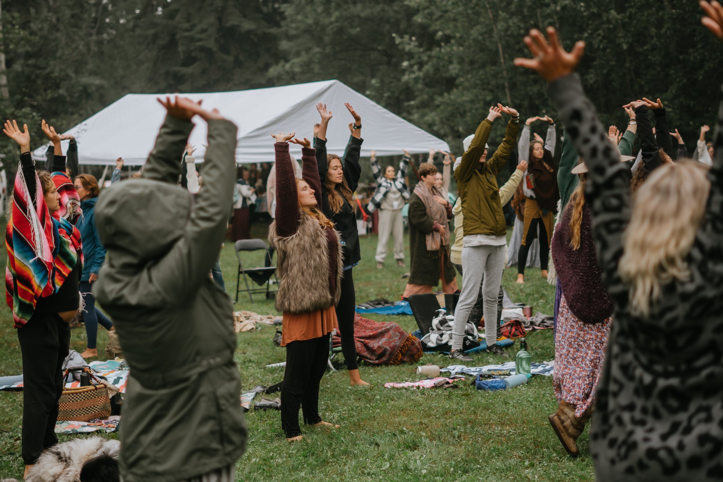 A group of people practicing outdoor yoga or meditation in a field with raised arms, surrounded by trees and a white tent. Participants are dressed in casual clothing, with some sitting or standing on blankets.