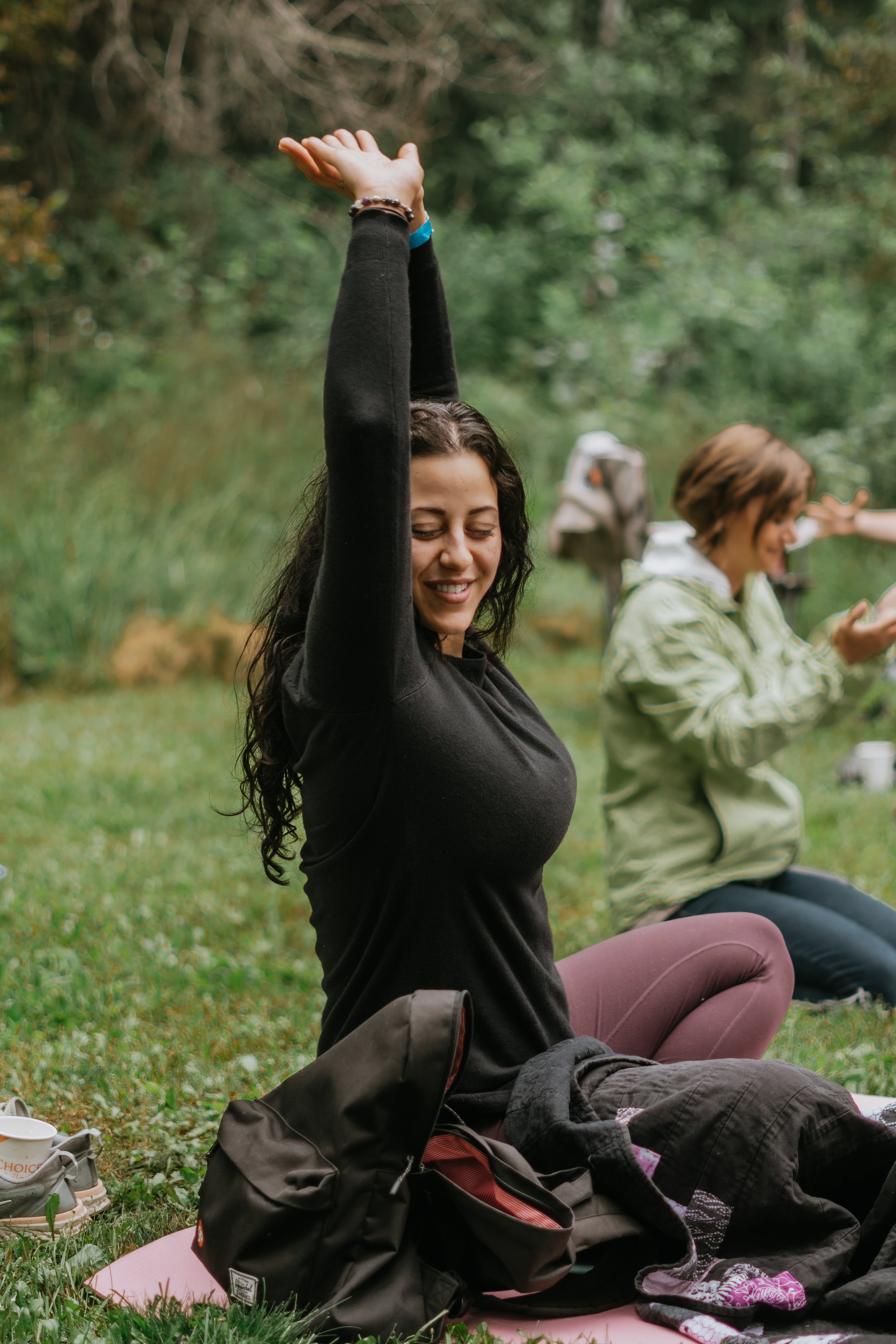 A woman practicing yoga outdoors, sitting on a yoga mat with her arms raised, wearing a black top and maroon leggings. A backpack and a blanket are beside her. Another person is in the background.