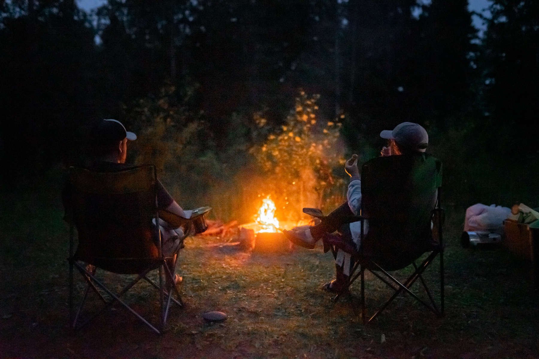 Two people sitting in camping chairs by a campfire at night, surrounded by trees.