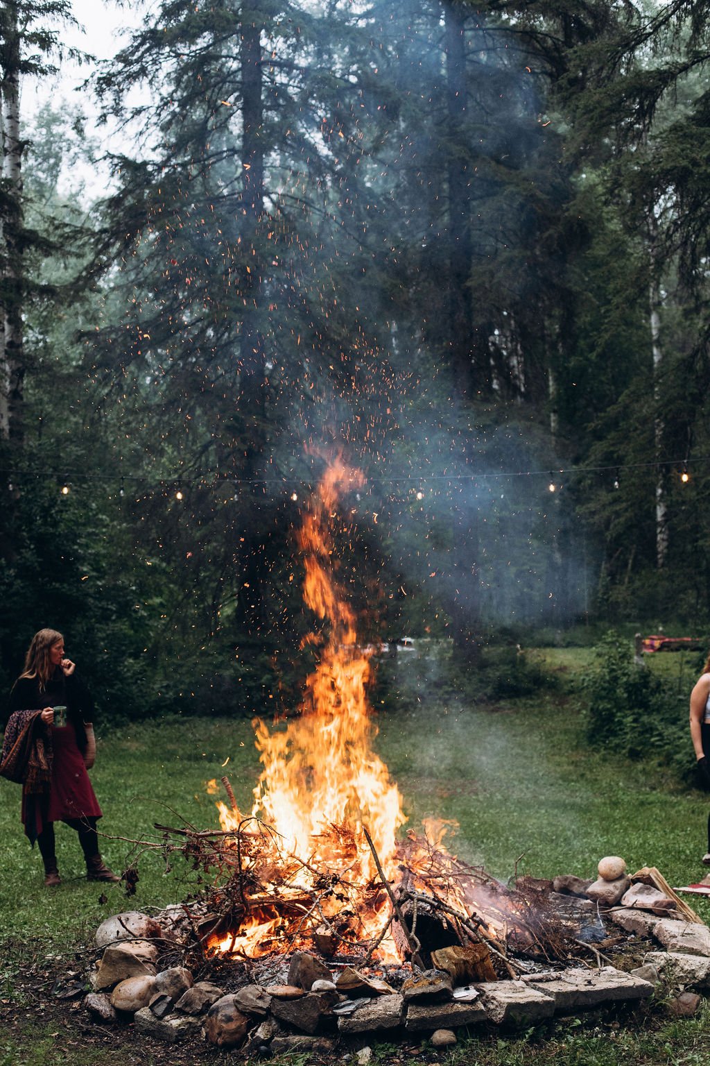Large bonfire with people in a forest setting, surrounded by trees, string lights, and stone fire ring.