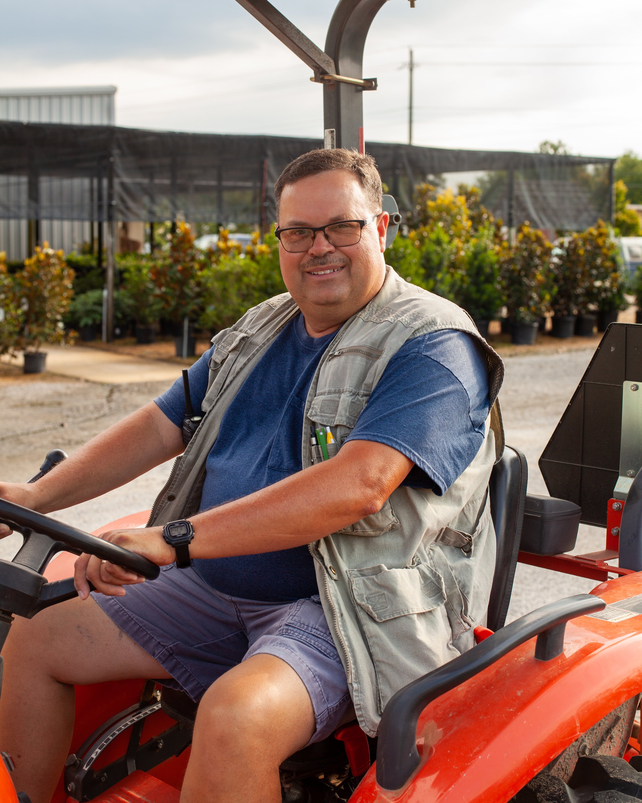 A man smiling while sitting on a tractor at a plant nursery, wearing glasses, a blue t-shirt, gray shorts, and a beige vest with pens in the pocket.