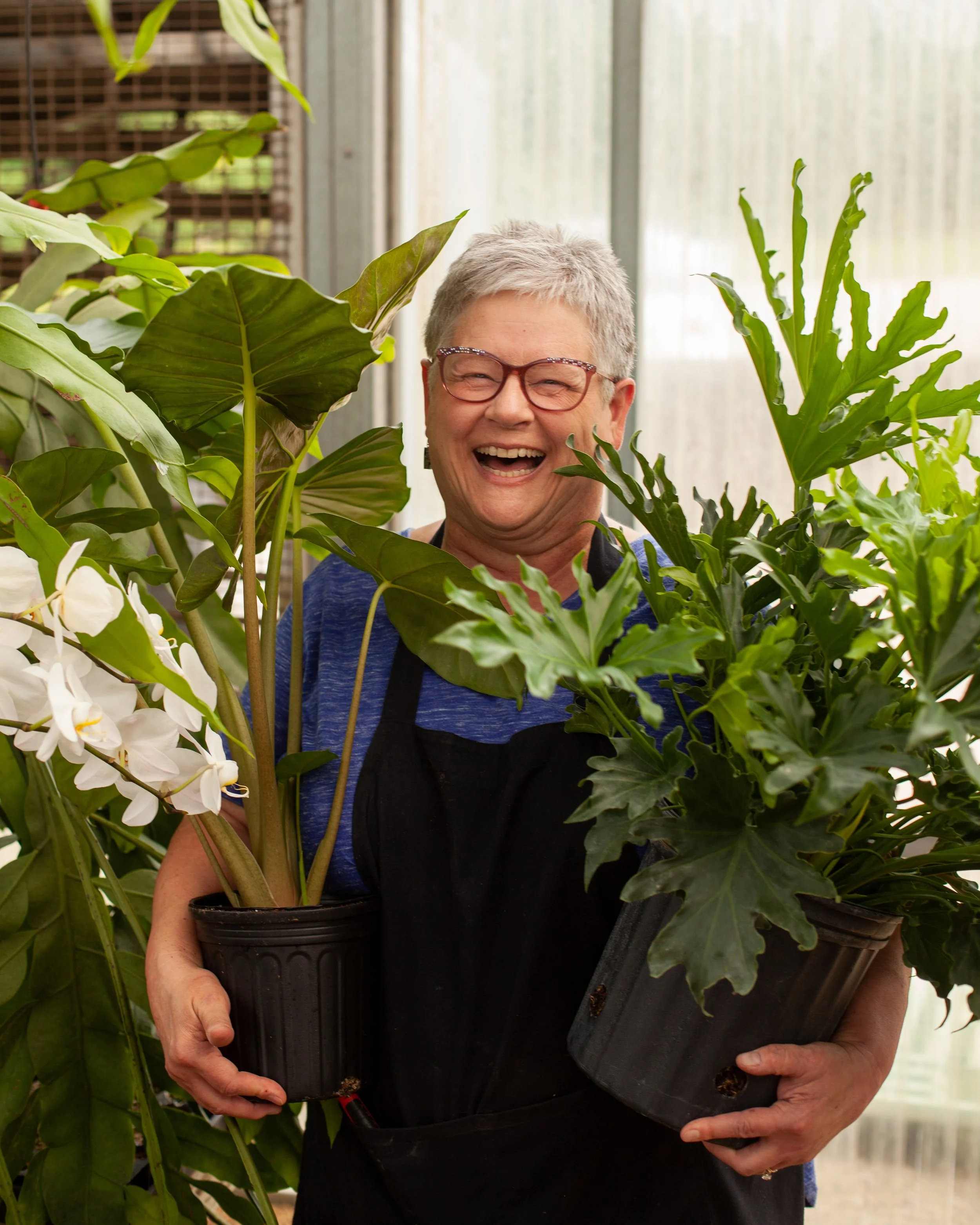 An older woman with short gray hair and glasses smiling while holding two potted plants.