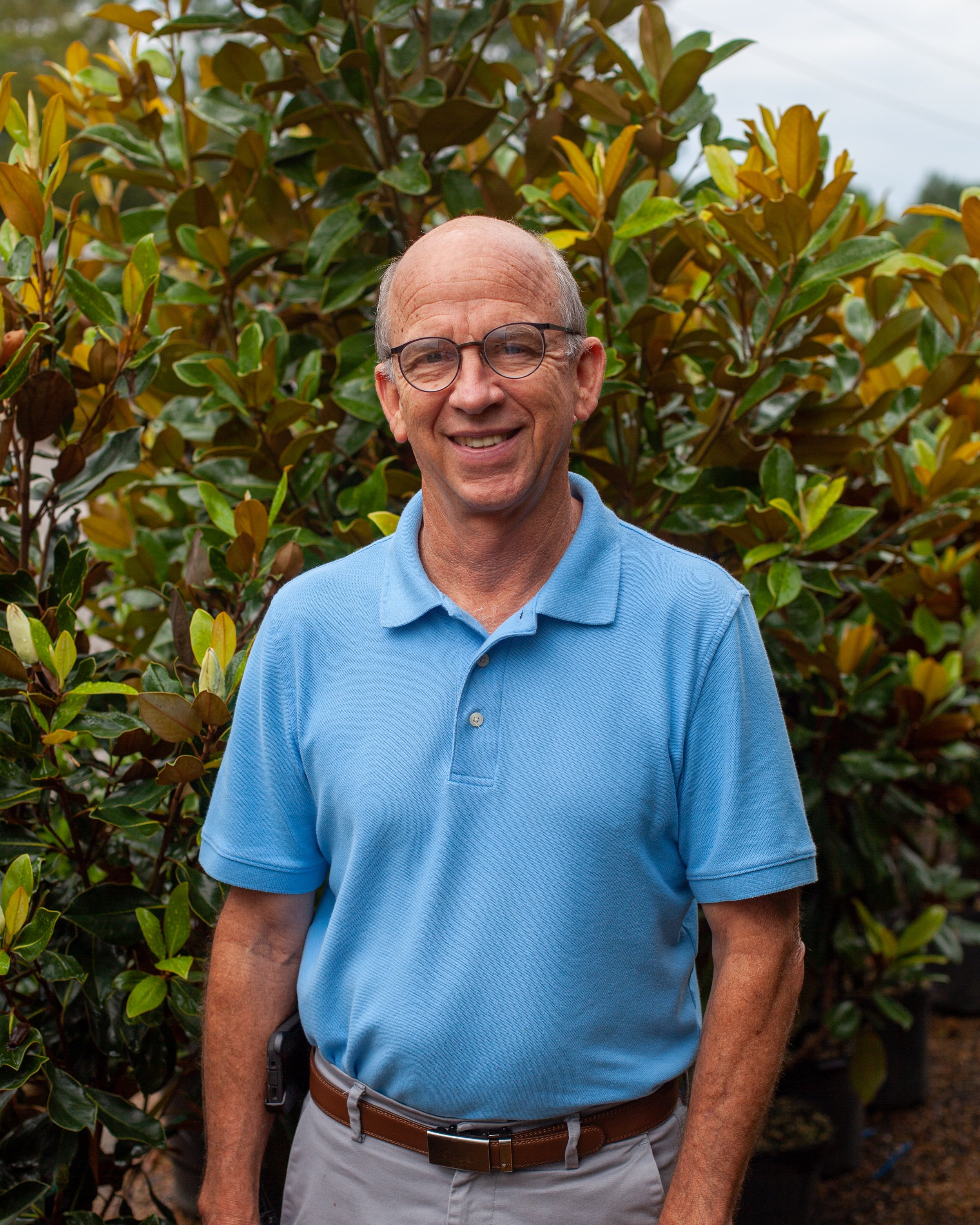 An older man wearing glasses and a blue polo shirt standing outdoors in front of a lush, green shrub.