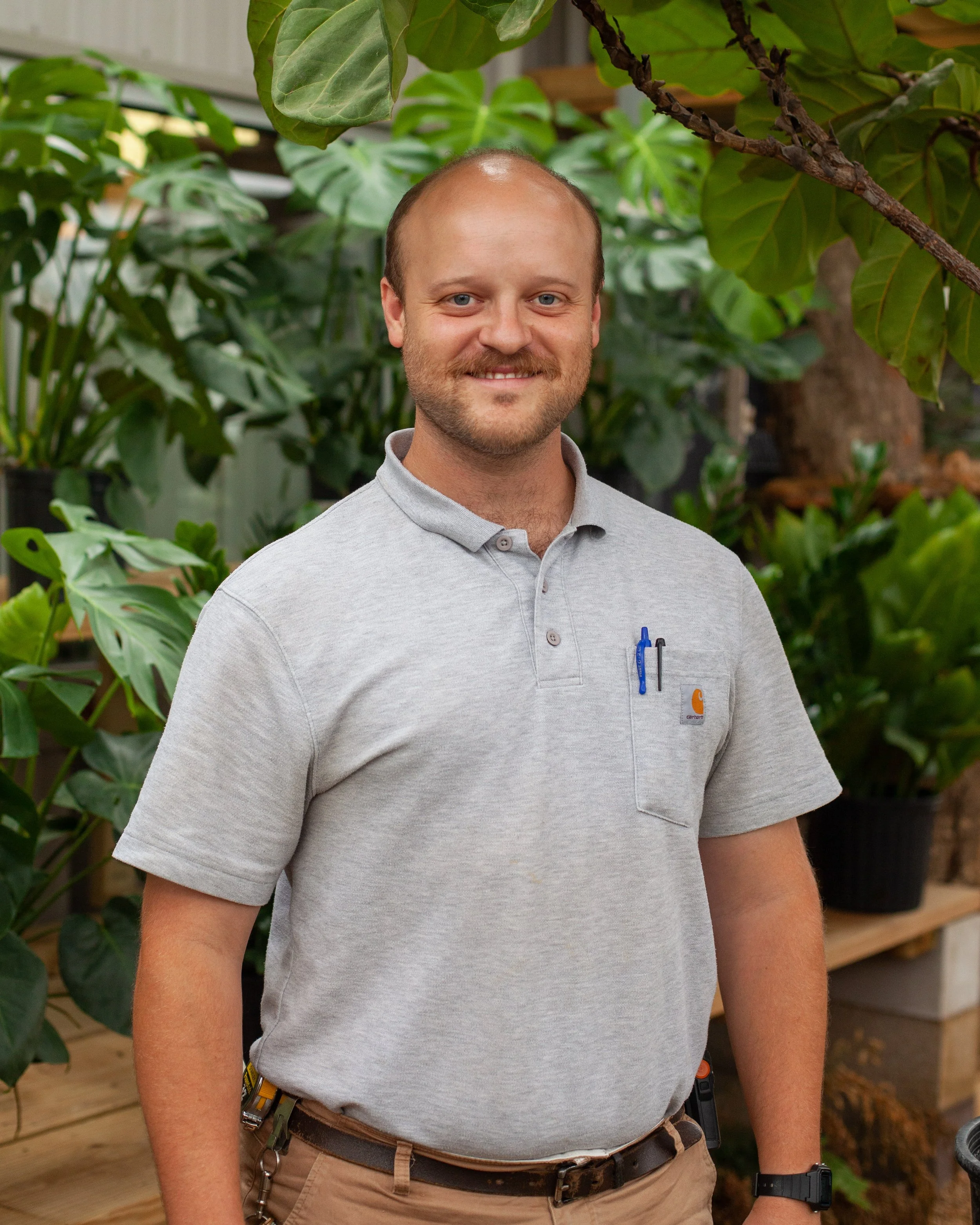 A man in a light gray polo shirt standing in a greenhouse with large green plants.