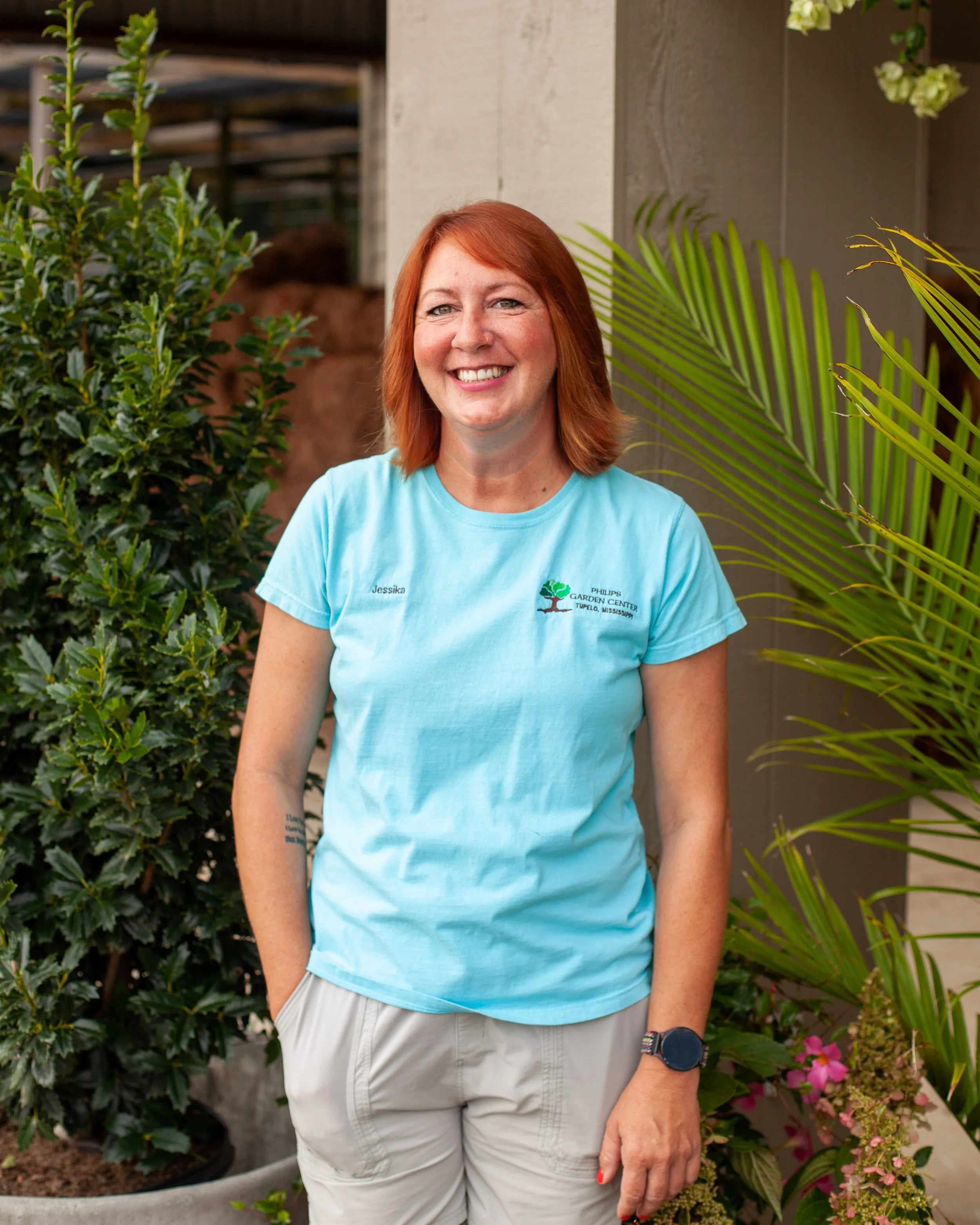 A woman with red hair, smiling, wearing a light blue t-shirt with a garden center logo, standing outdoors near greenery and plants.
