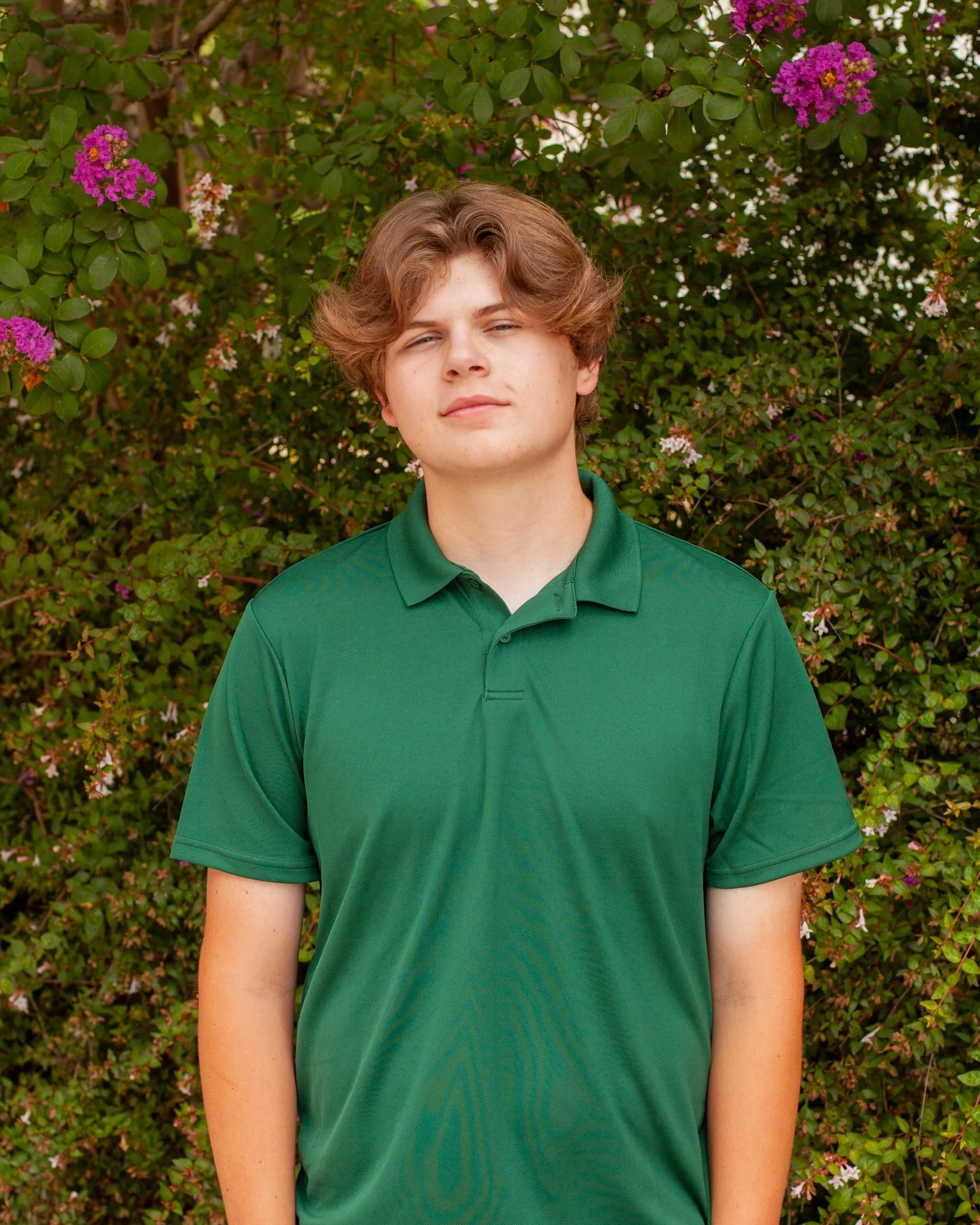 Young man with light brown hair wearing a green polo shirt, standing in front of a bush with purple and pink flowers.