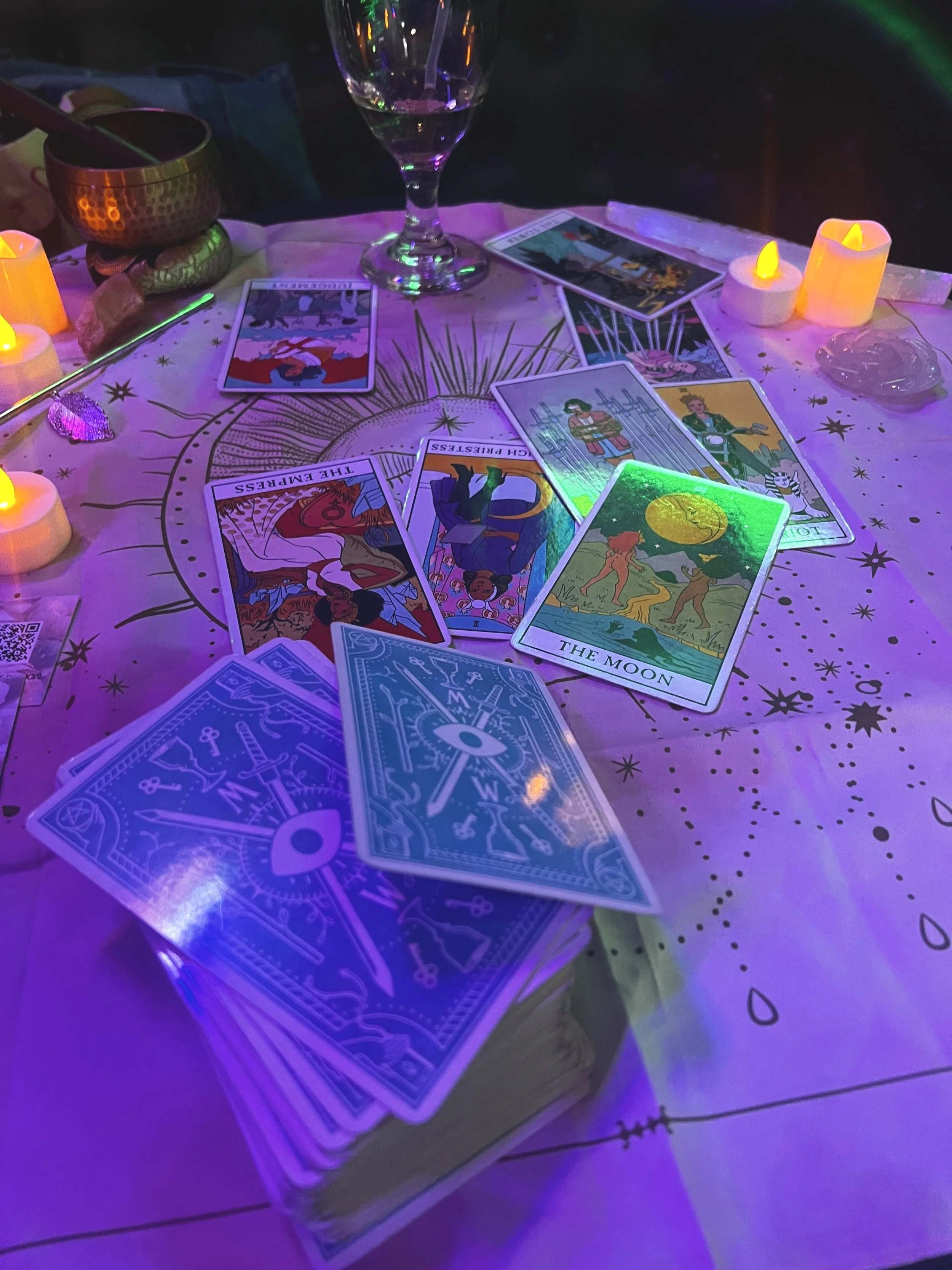 A tarot card reading setup on a table with lit candles, a glass of drink, tarot cards spread out, and decorative cloth under purple lighting.