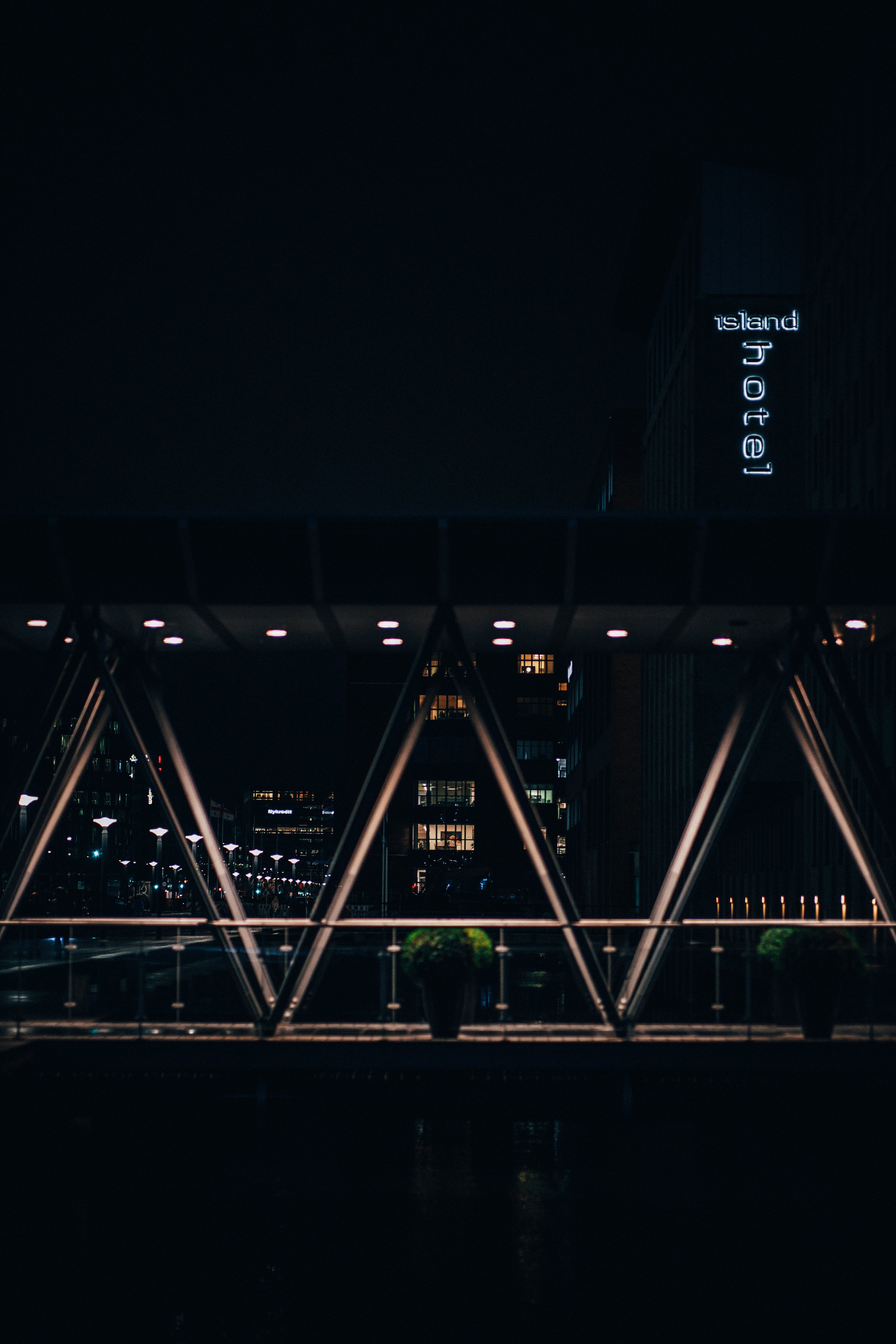 Night view of a modern bridge with illuminated metal framework, Potted plants on the bridge, city buildings in the background, and a lit-up sign reading 'Island Hotel' on a tall building.