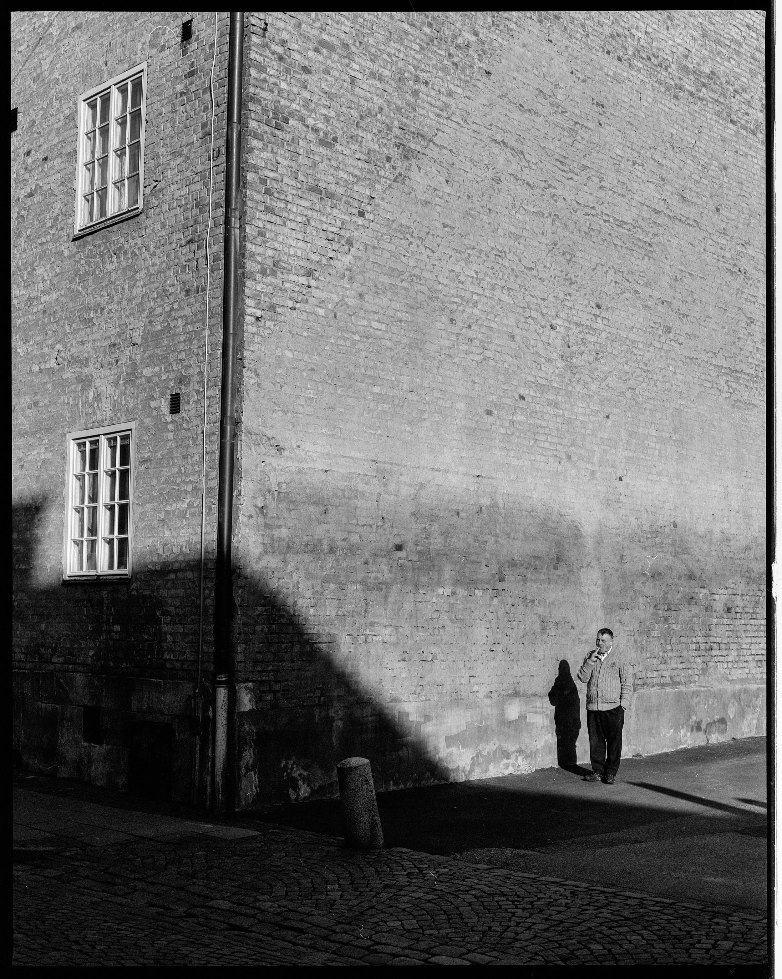 A black and white photo of a man standing on a corner, talking on his phone, cast in shadow on a tall brick building wall.