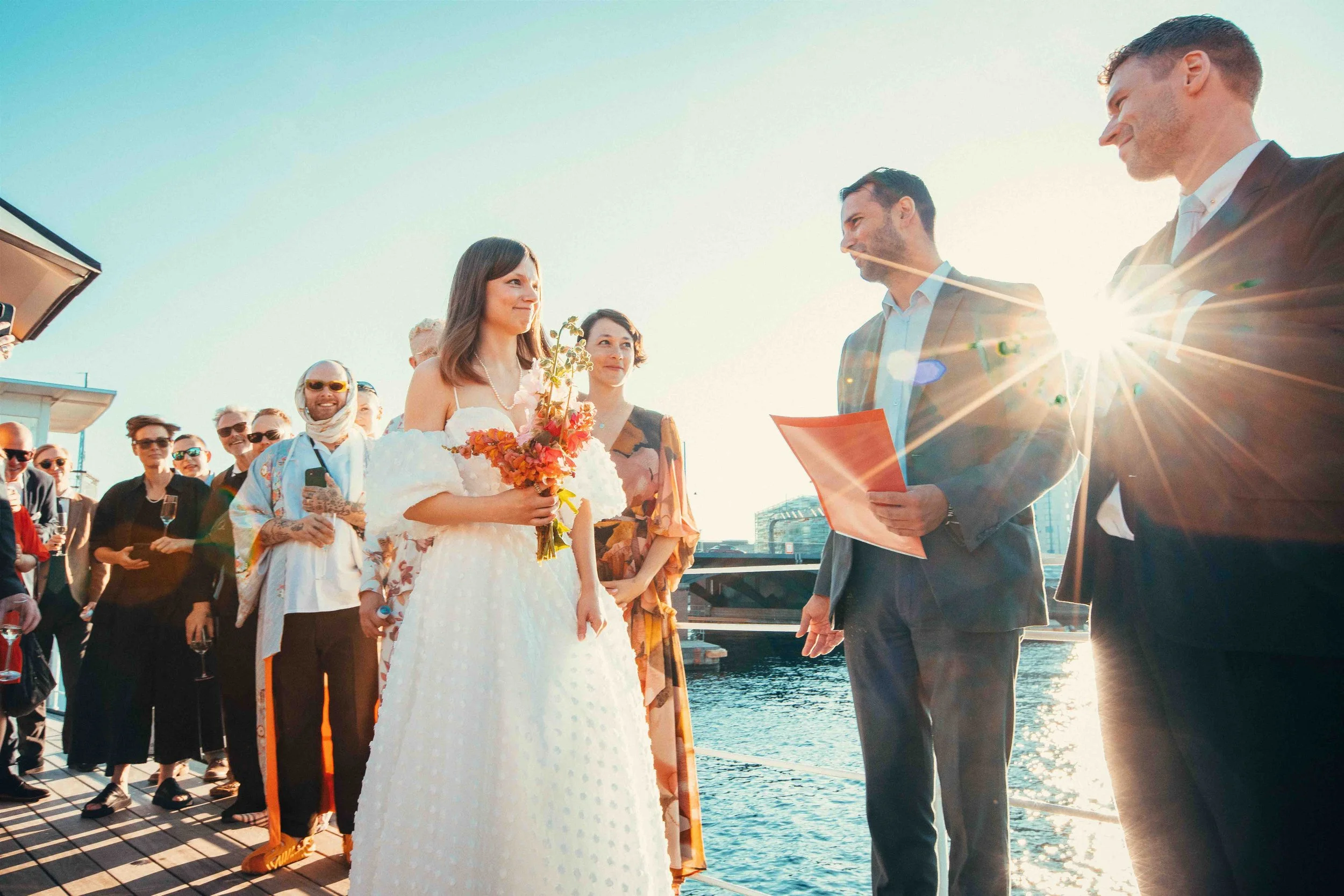 A wedding ceremony takes place on a dock by the water, with a bride in a white dress holding a bouquet of flowers and men in suits officiating, as sun rays create a backlit effect.
