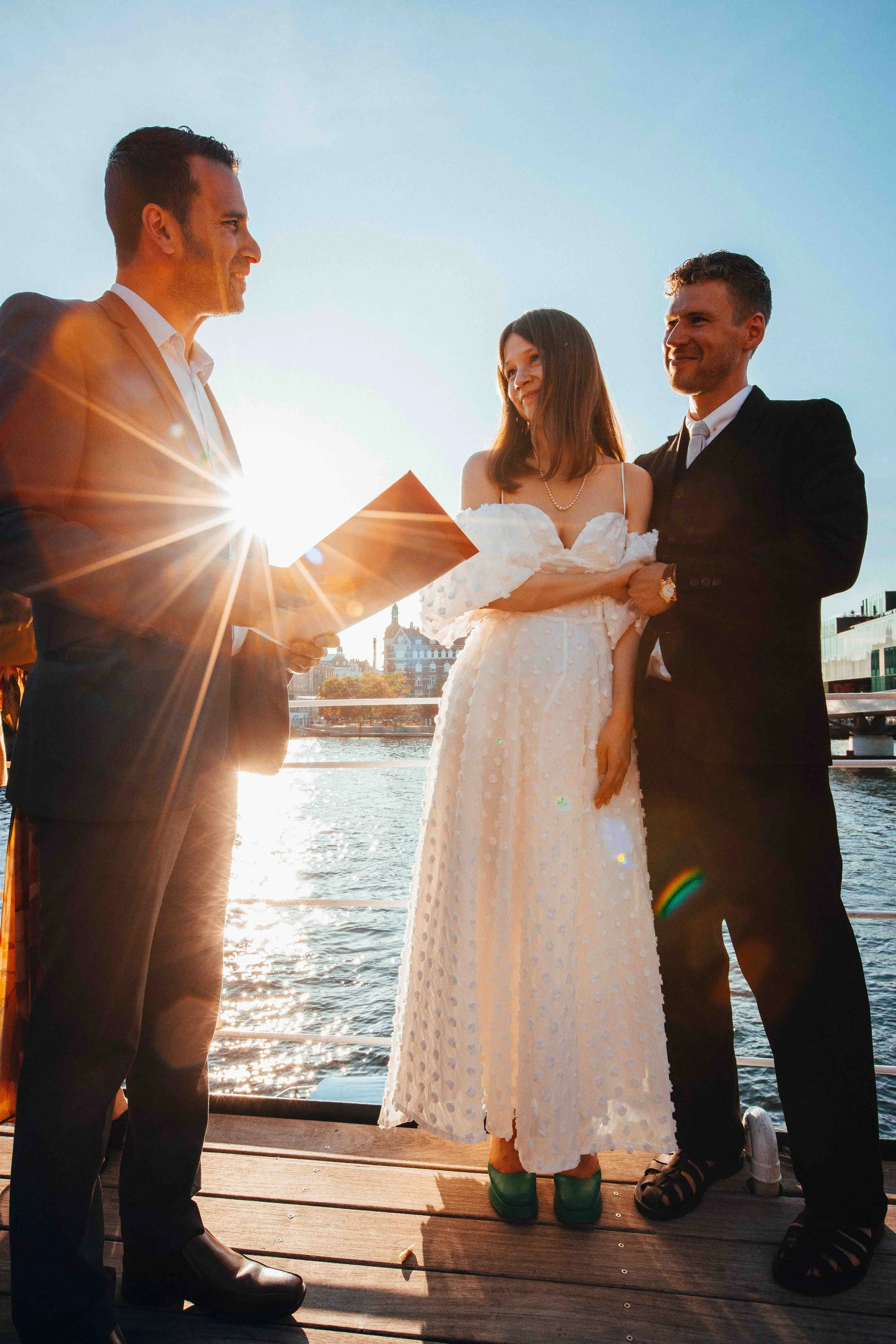 A wedding ceremony outdoors by the water at sunset with three people: a man reading from a book, a woman in a wedding dress, and a man in a black suit. The sun creates a starburst effect and lens flare.