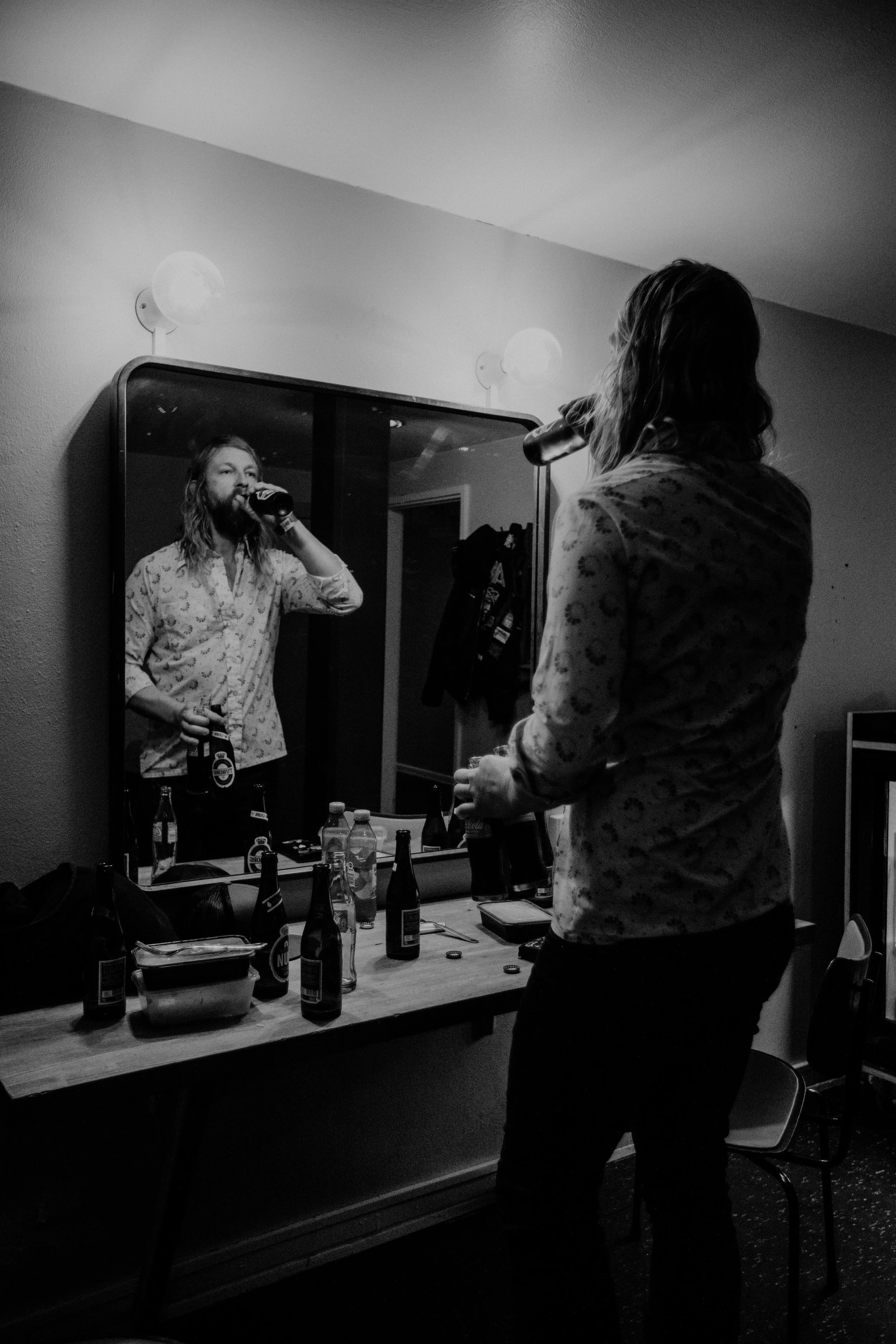 A woman with shoulder-length hair, wearing a patterned shirt, is standing in front of a mirror, drinking from a bottle. Her reflection is visible in the mirror, and the scene includes a table with several bottles and cans, some food containers, and a