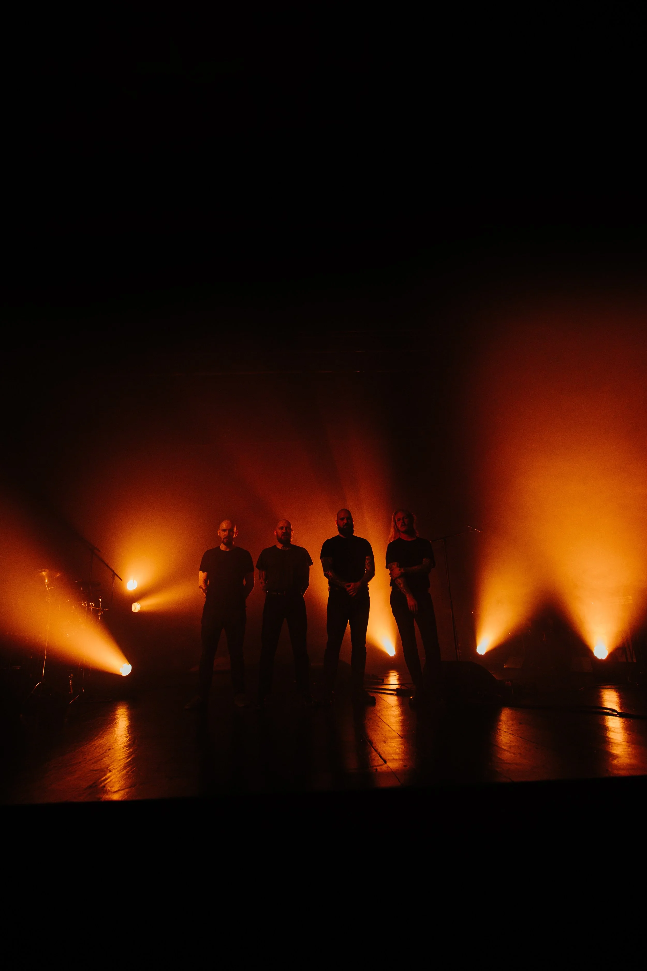 Four silhouetted men on stage with orange backlighting and fog, possibly during a concert or performance.