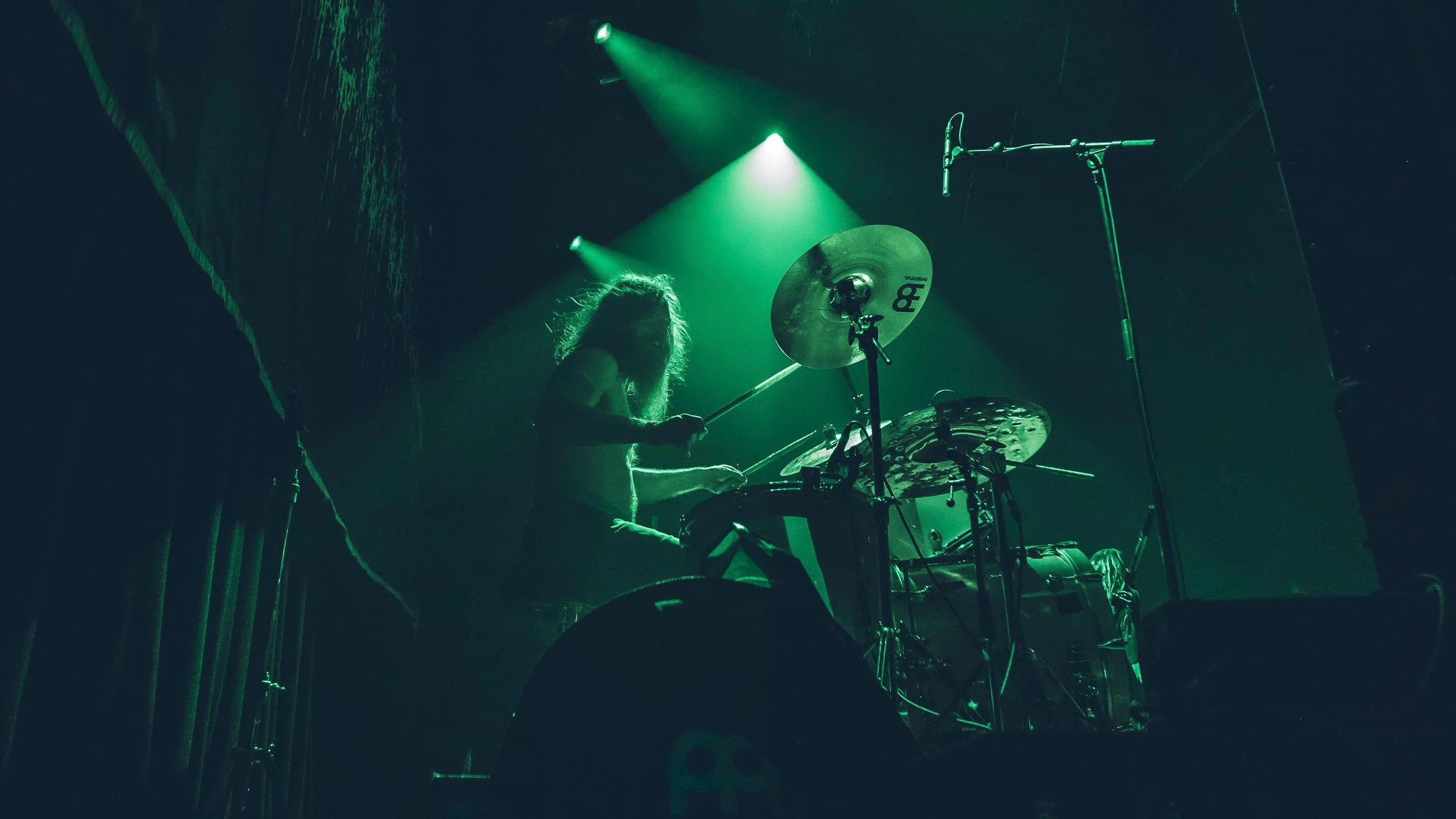A female drummer playing on stage under green spotlight, surrounded by drums and cymbals.