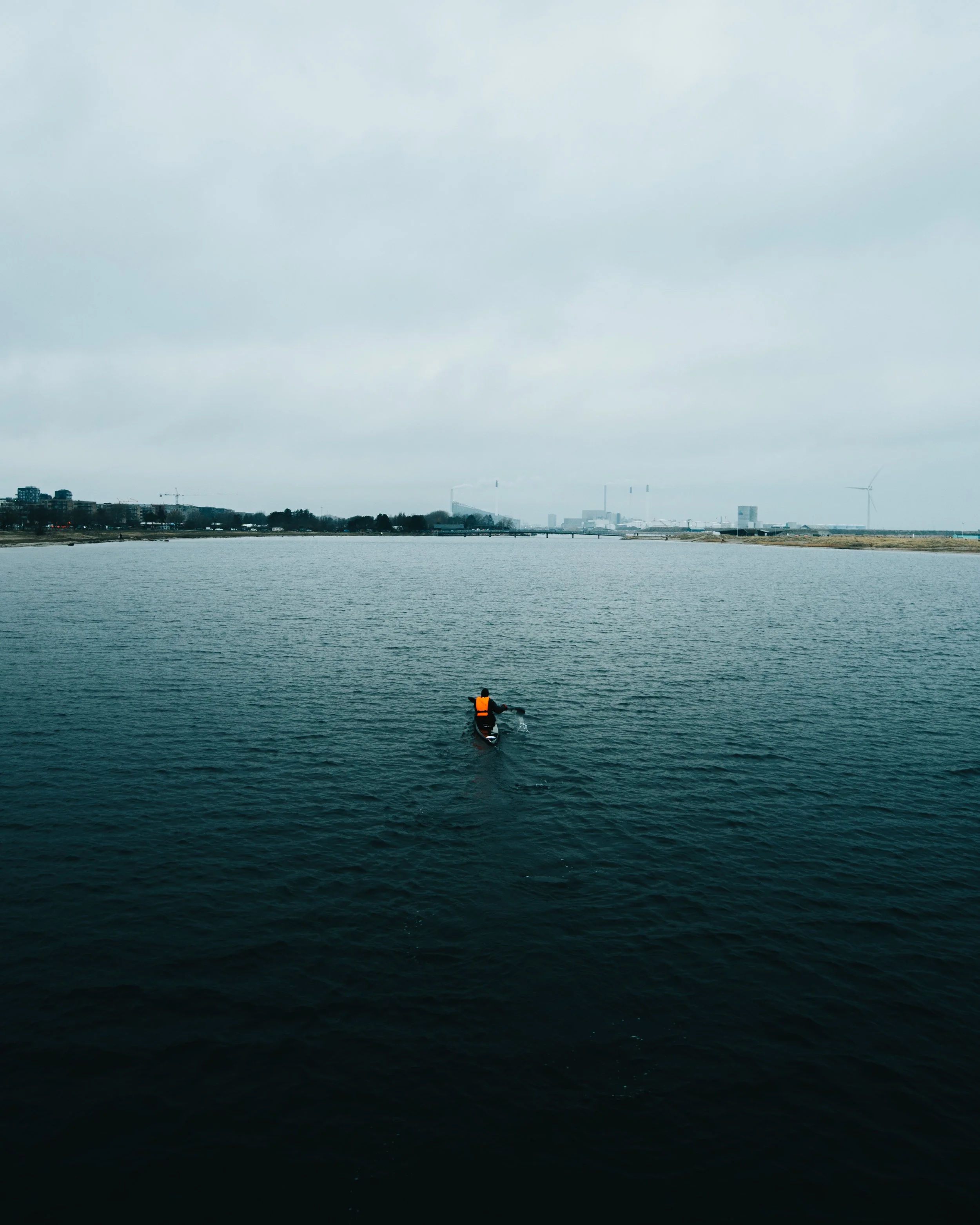 A person in an orange life vest kayaking on a large body of water on a cloudy day, with city buildings and a wind turbine visible in the distance.