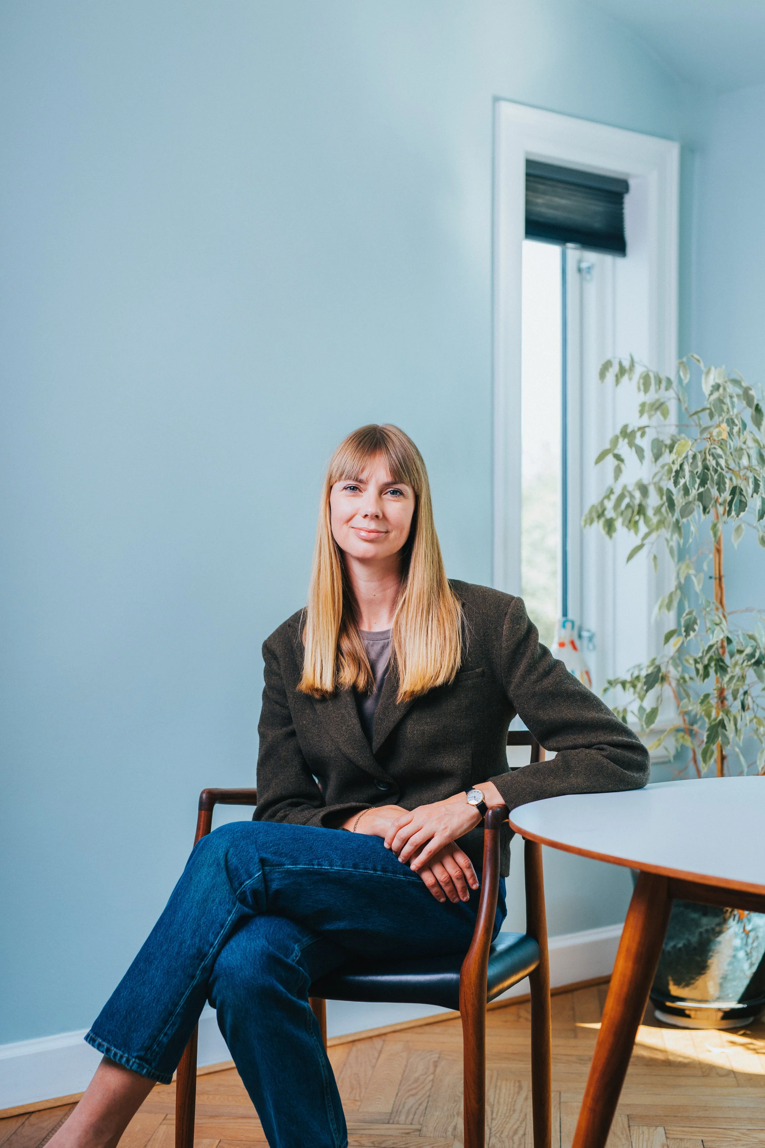 A woman with long blond hair sitting in a chair by a window in a room with light blue walls and wooden flooring.