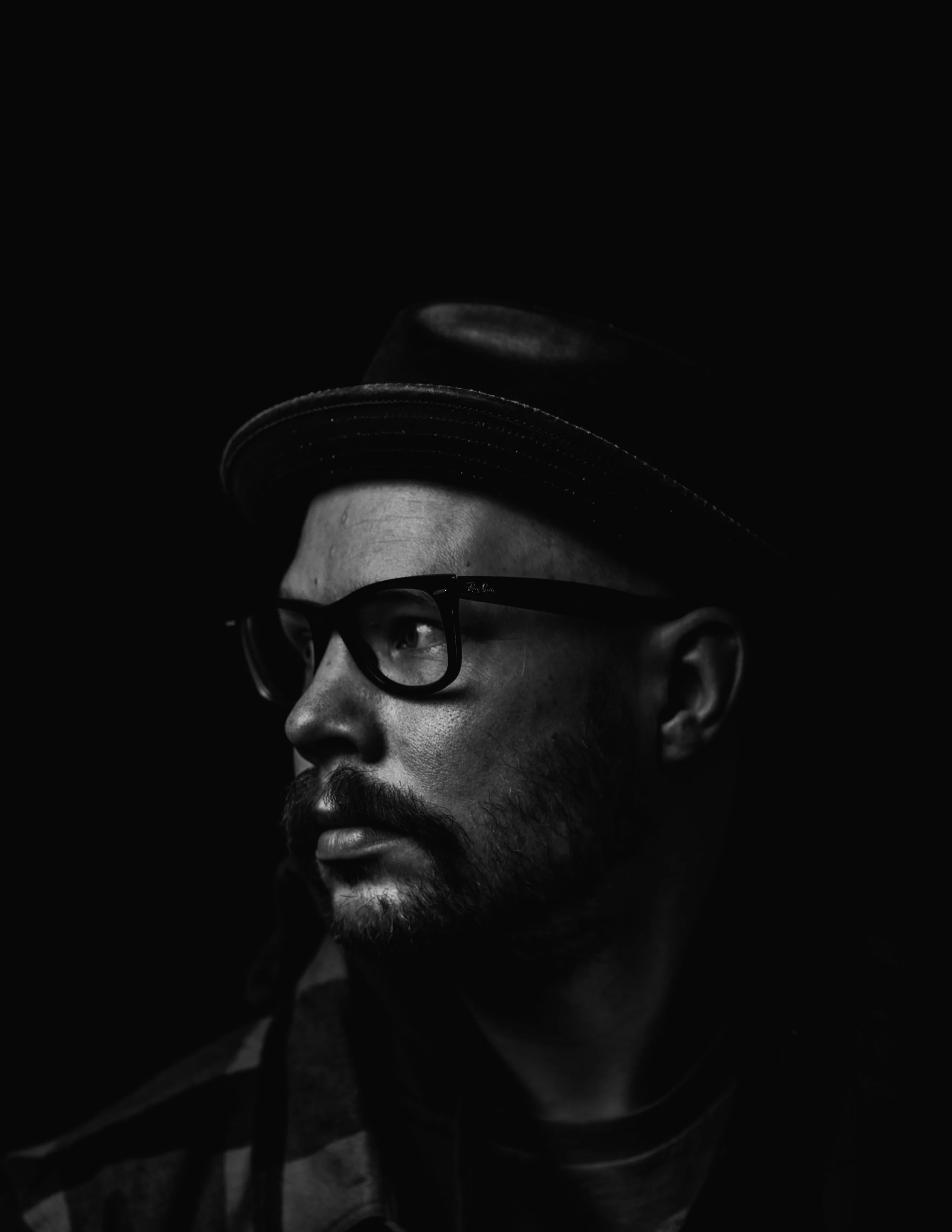 Black and white portrait of a man wearing a hat, glasses, and a striped shirt, looking to the side against a dark background.