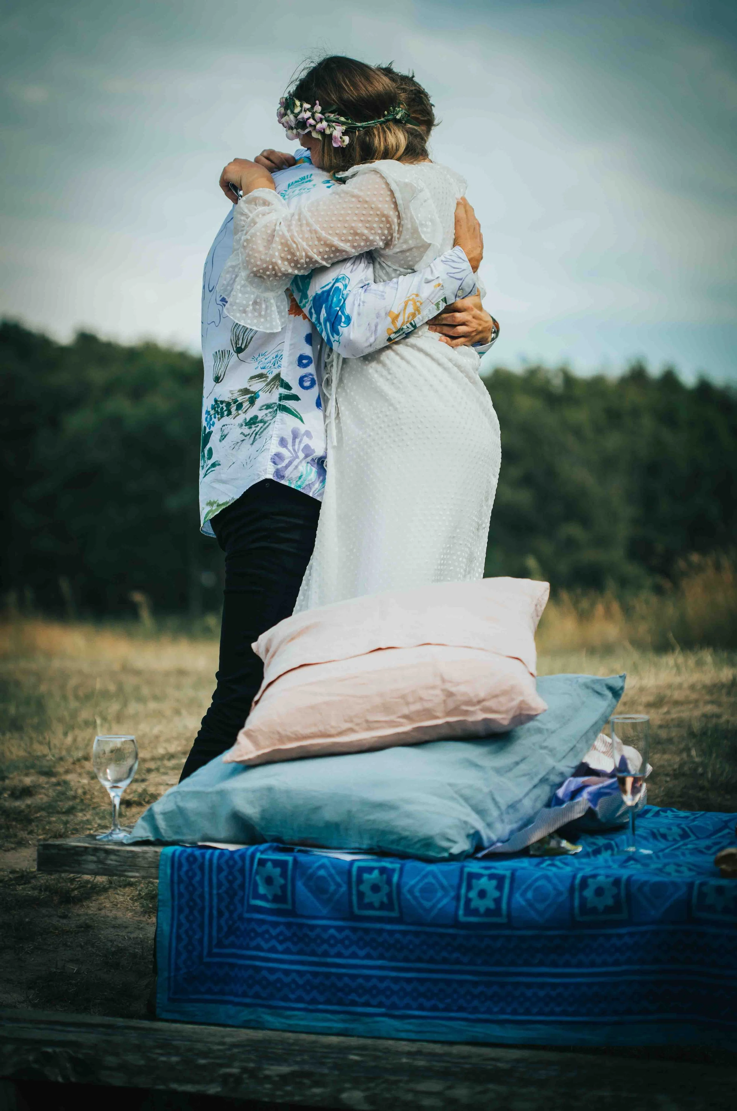 A couple embracing during an outdoor picnic on a blanket with pillows and glasses of wine, set against a grassy field and cloudy sky.