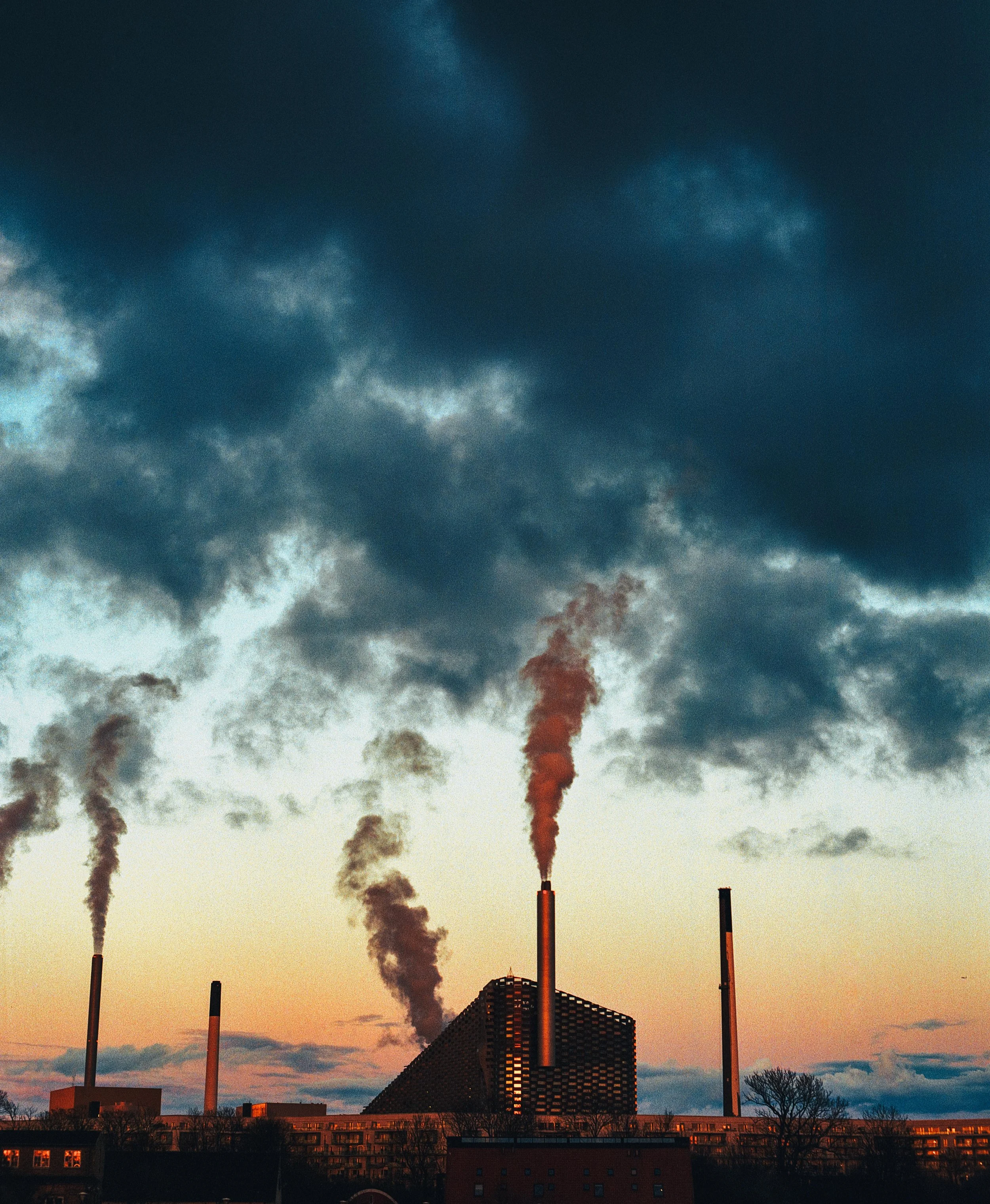 An industrial power plant with multiple smokestacks emitting dark smoke during sunset, with a sky filled with clouds.