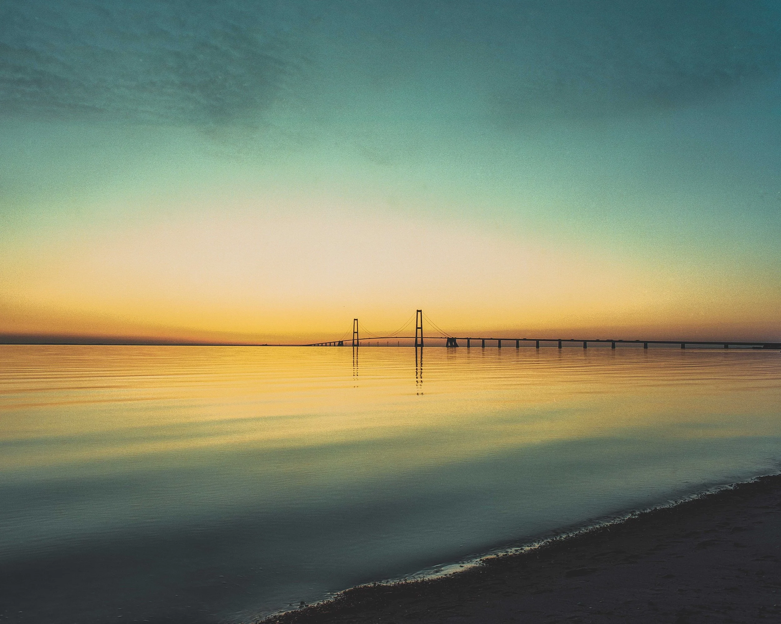 A bridge over calm water during a sunset or sunrise, with the sky and water reflecting soft colors.