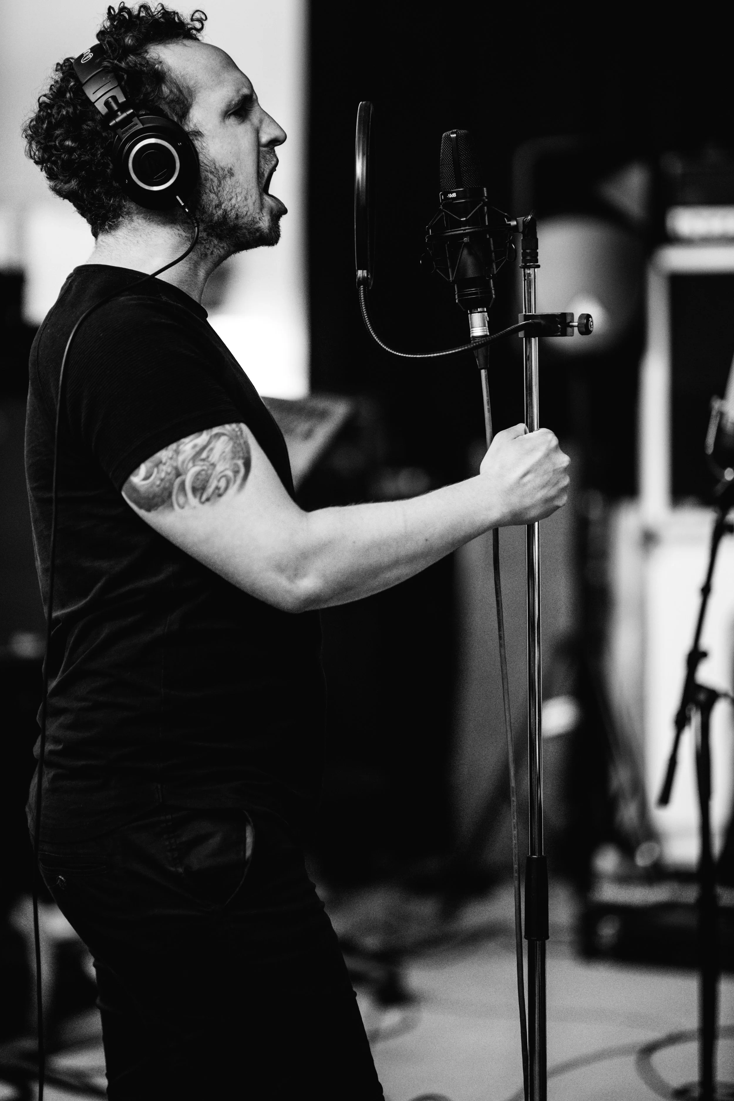 A man singing passionately into a microphone in a recording studio, wearing headphones and a black t-shirt, with tattoos on his arm, in a black-and-white photograph.
