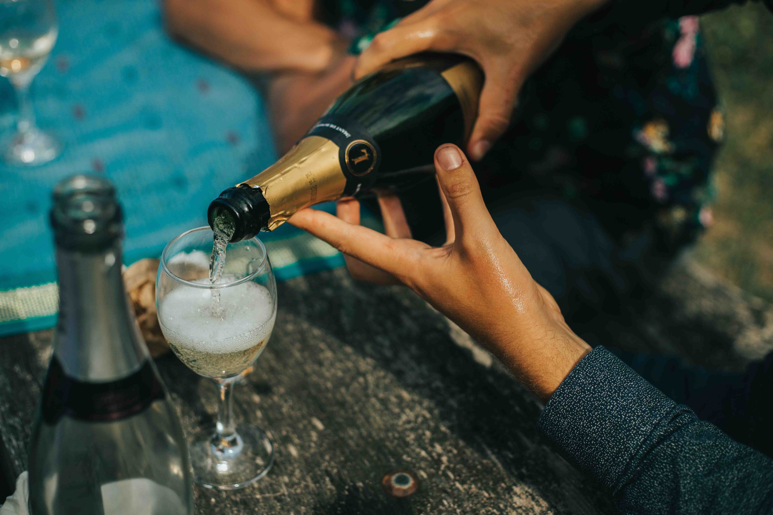 Person pouring champagne into a glass outdoors on a picnic table.