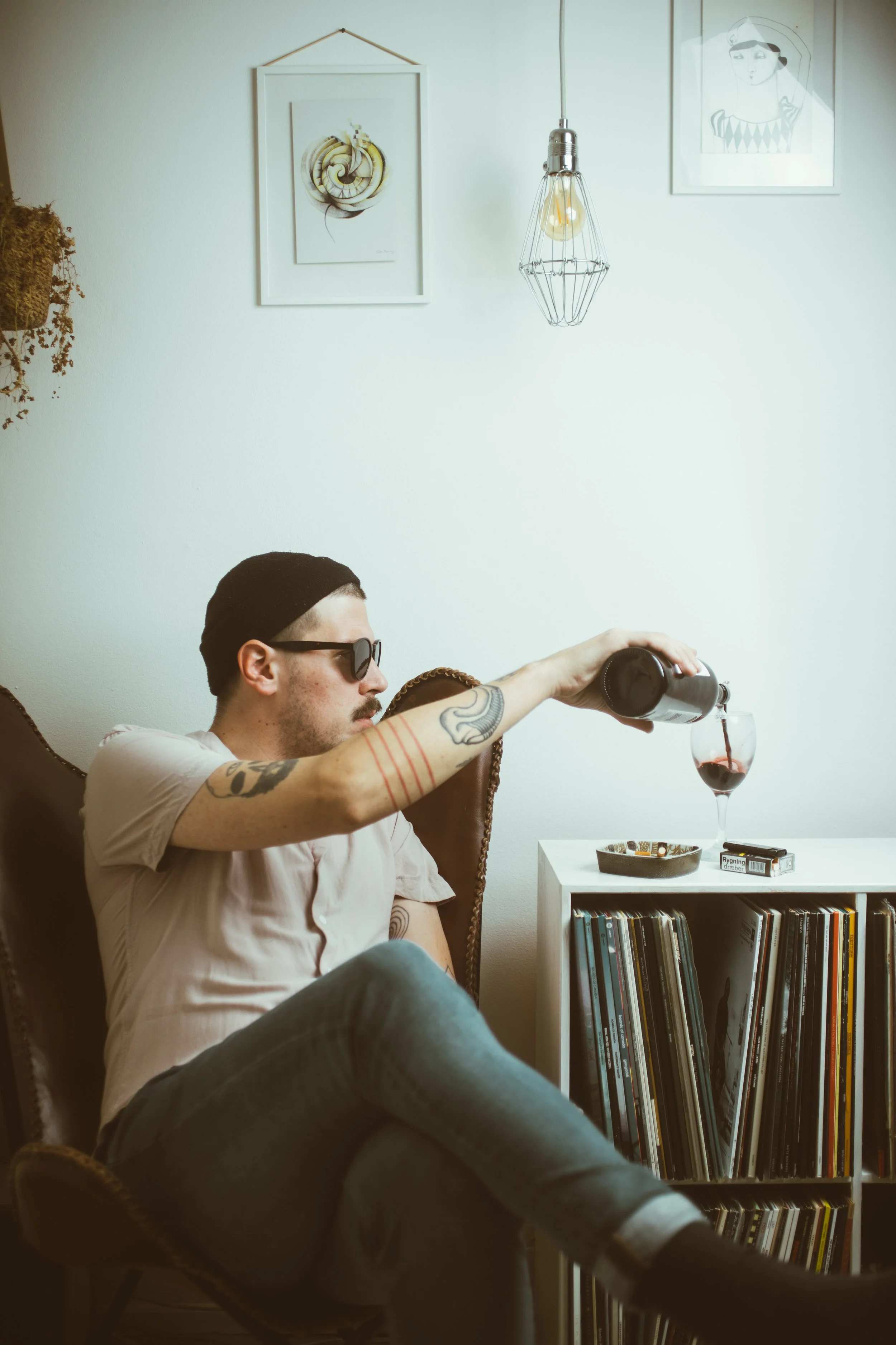 A man wearing sunglasses and a black beanie pours red wine into a glass while sitting on a vintage armchair beside a white bookshelf filled with records. The room has minimalist decor with framed artwork and a hanging light fixture on the wall.