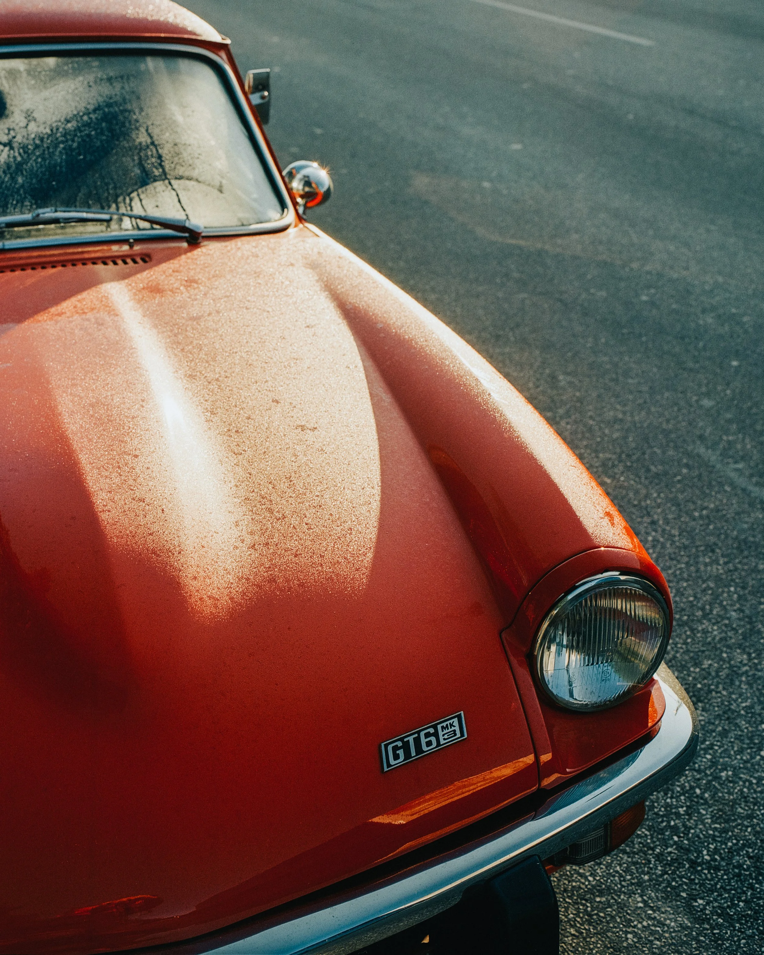 Close-up of a vintage red Pontiac GT6 car with a dusty hood, headlight, and a GT6 badge, parked on asphalt.