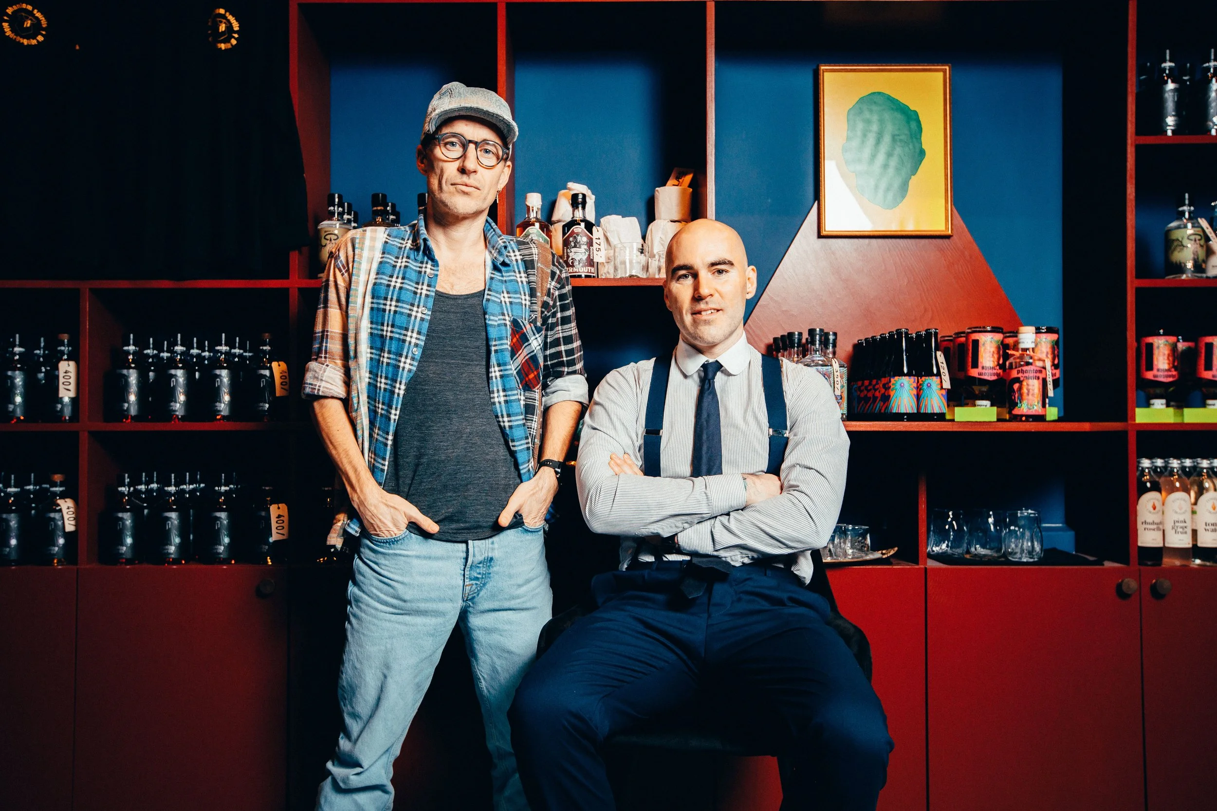 Two men pose in front of a red and blue shelving unit stocked with bottles and accessories. The man on the left stands with hands in pockets, wearing glasses, a cap, a plaid shirt over a dark t-shirt, and jeans. The man on the right is seated, arms c