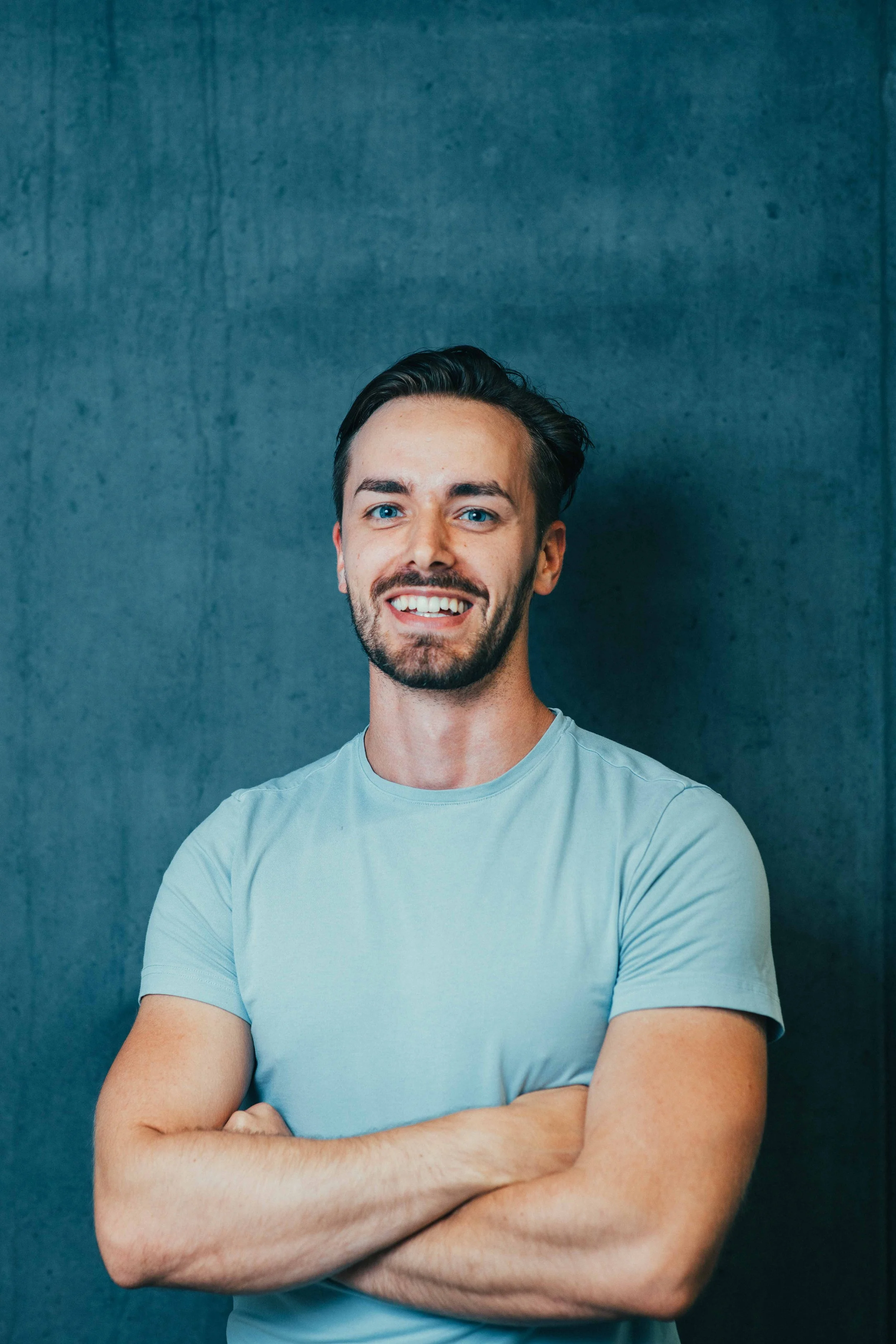 A young man with dark hair and a beard, smiling and standing with arms crossed in front of a textured gray wall.