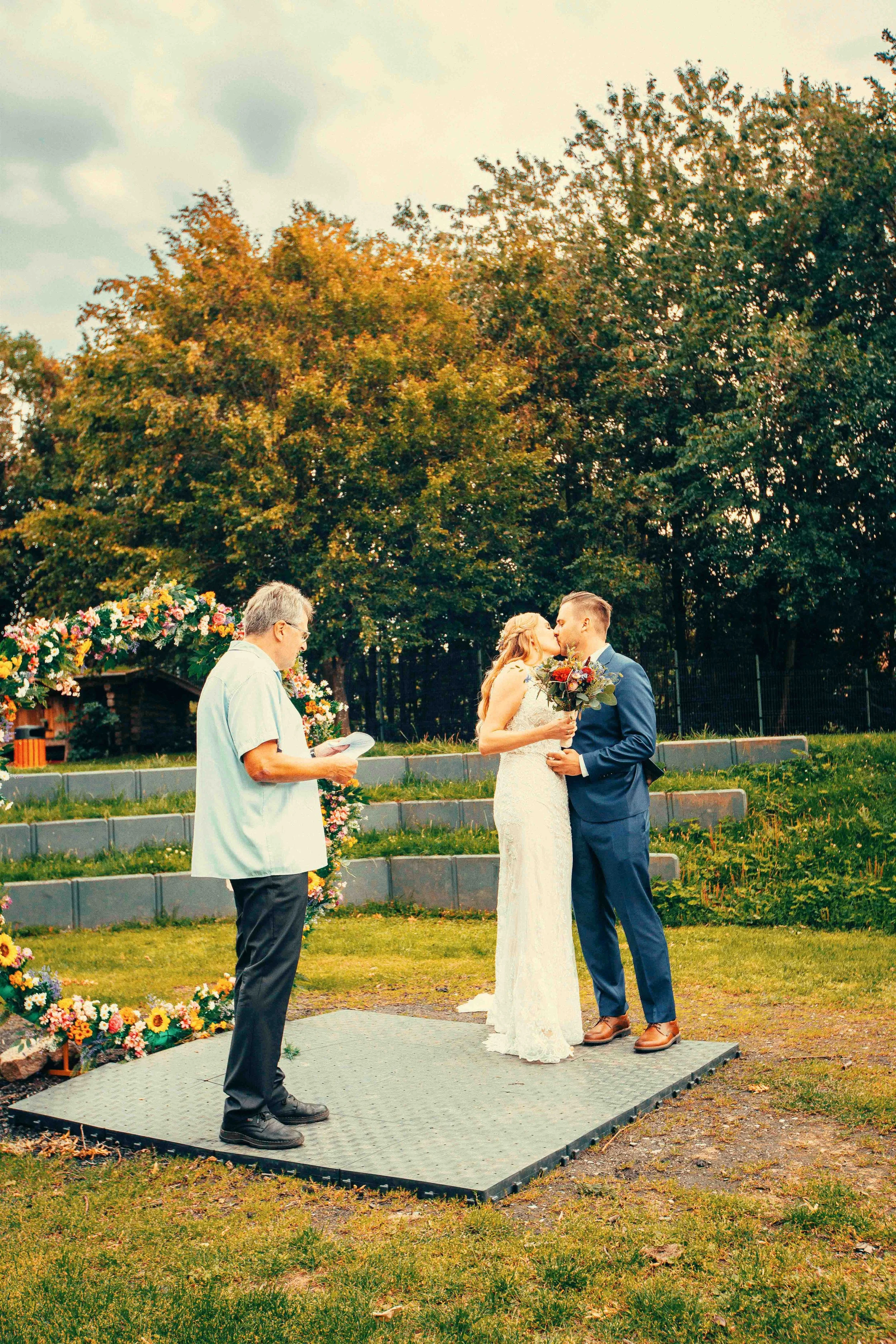A couple sharing a kiss during their outdoor wedding ceremony, with the officiant reading from a paper, flowers, and trees in the background.