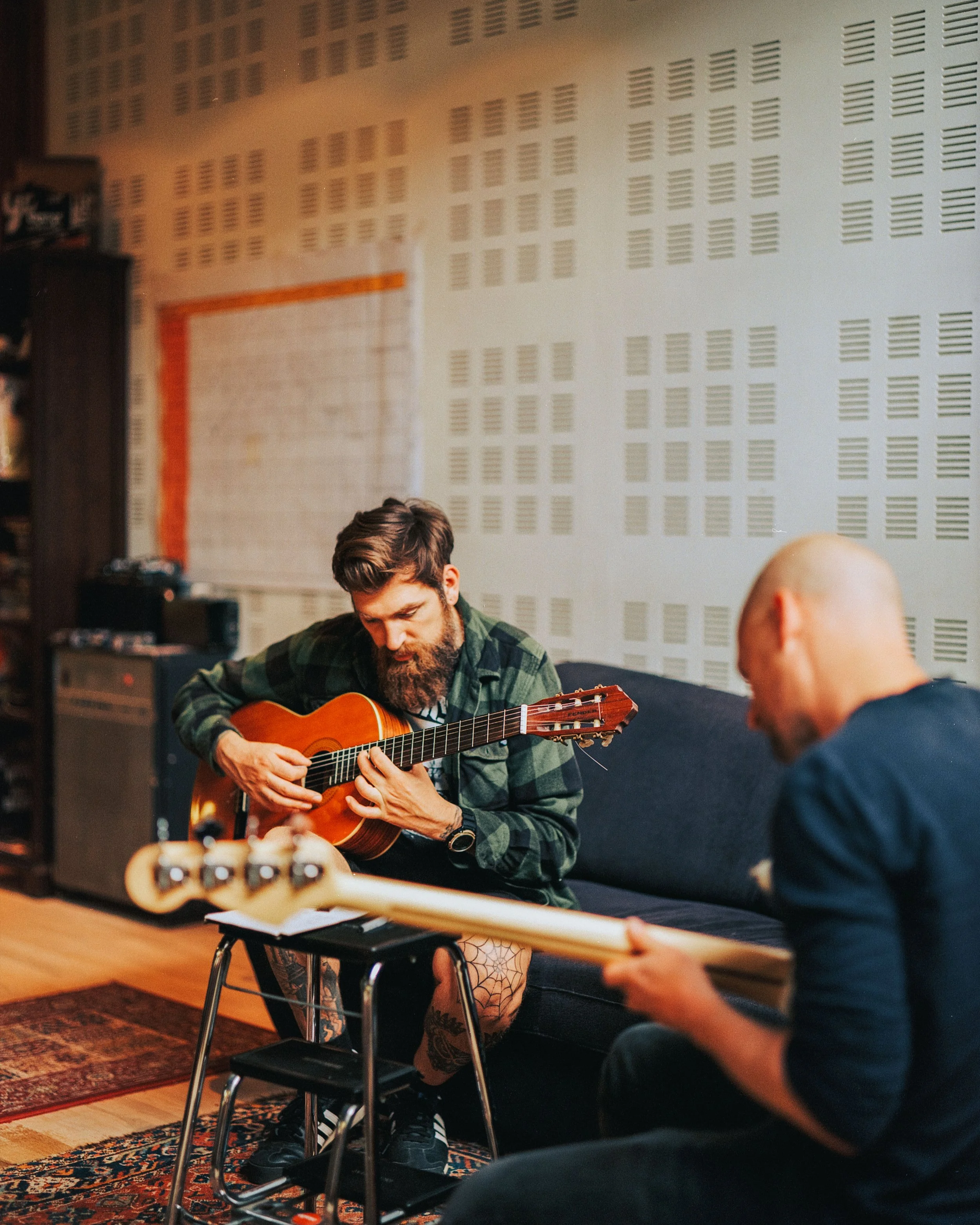 Two men playing acoustic guitar and bass guitar in a music studio, with soundproofing panels on the wall.