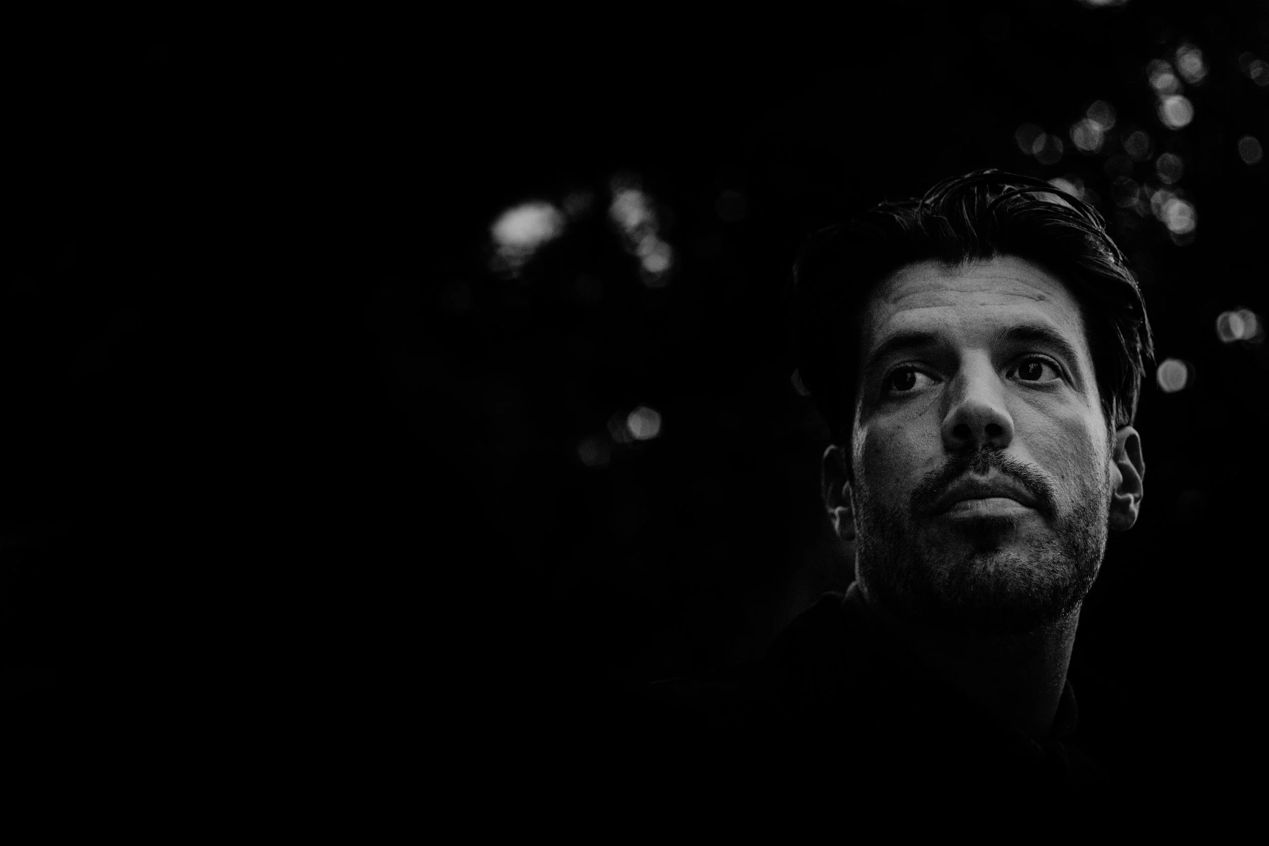 Black and white photograph of a man with a serious expression, looking off to the right, with a blurred background of trees and lights.