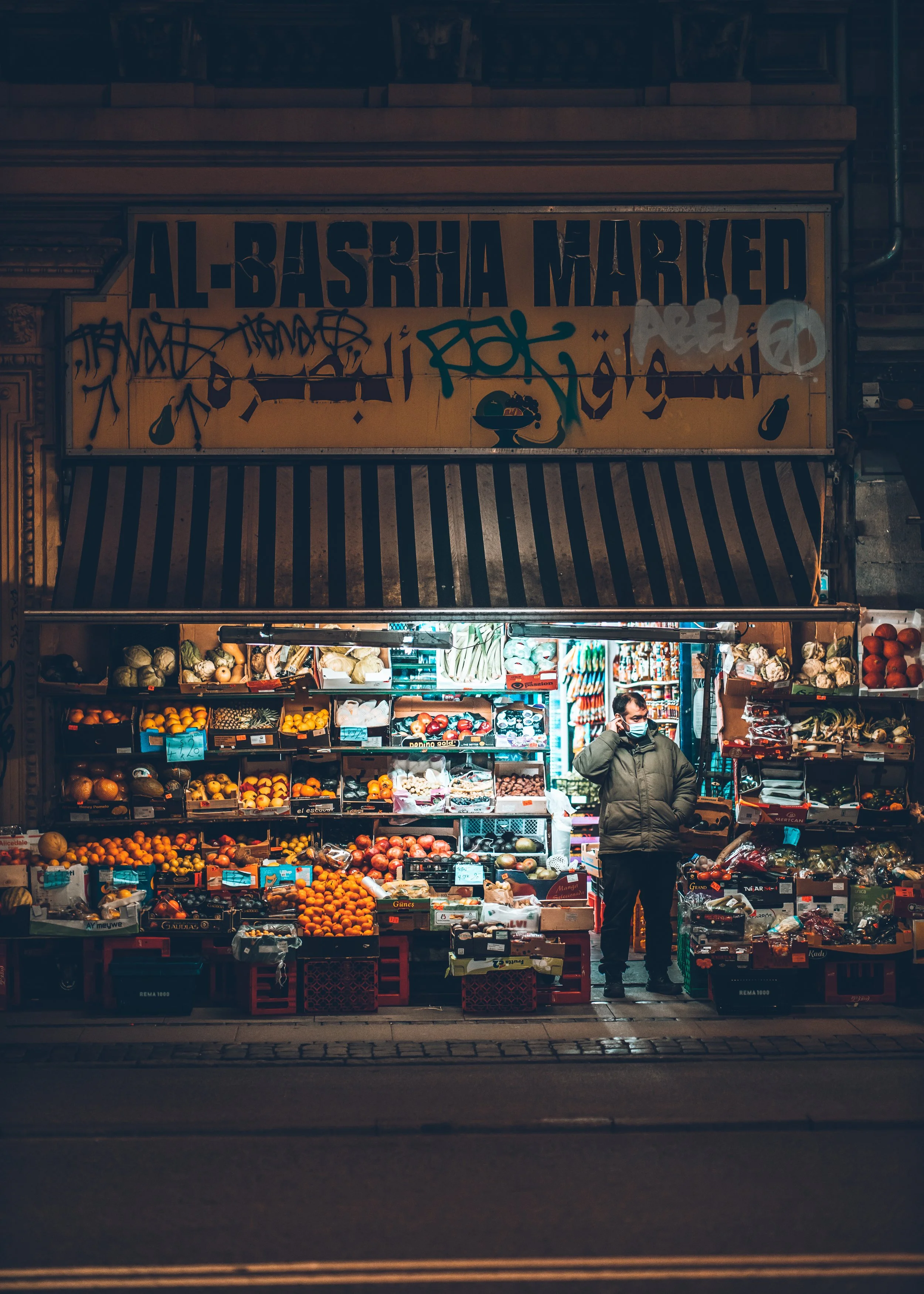 Nighttime street scene of a small fruit and vegetable stand with a man wearing a face mask, surrounded by colorful produce, in front of a building with a sign that reads 'Al-Basra Marked' and graffiti.