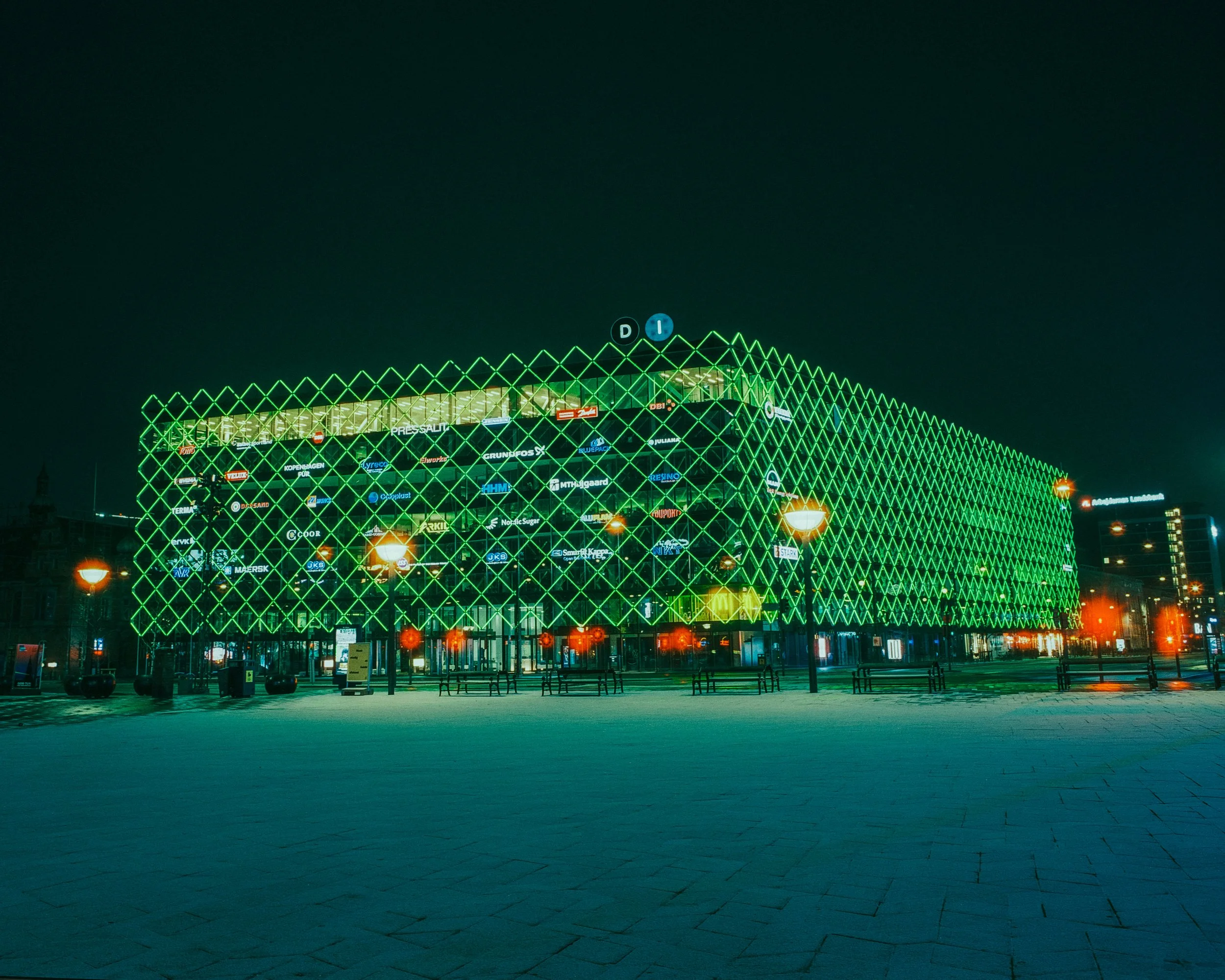 Night view of a modern building with a green, illuminated diamond-shaped lattice facade and a digital advertising display, surrounded by streetlights and benches.