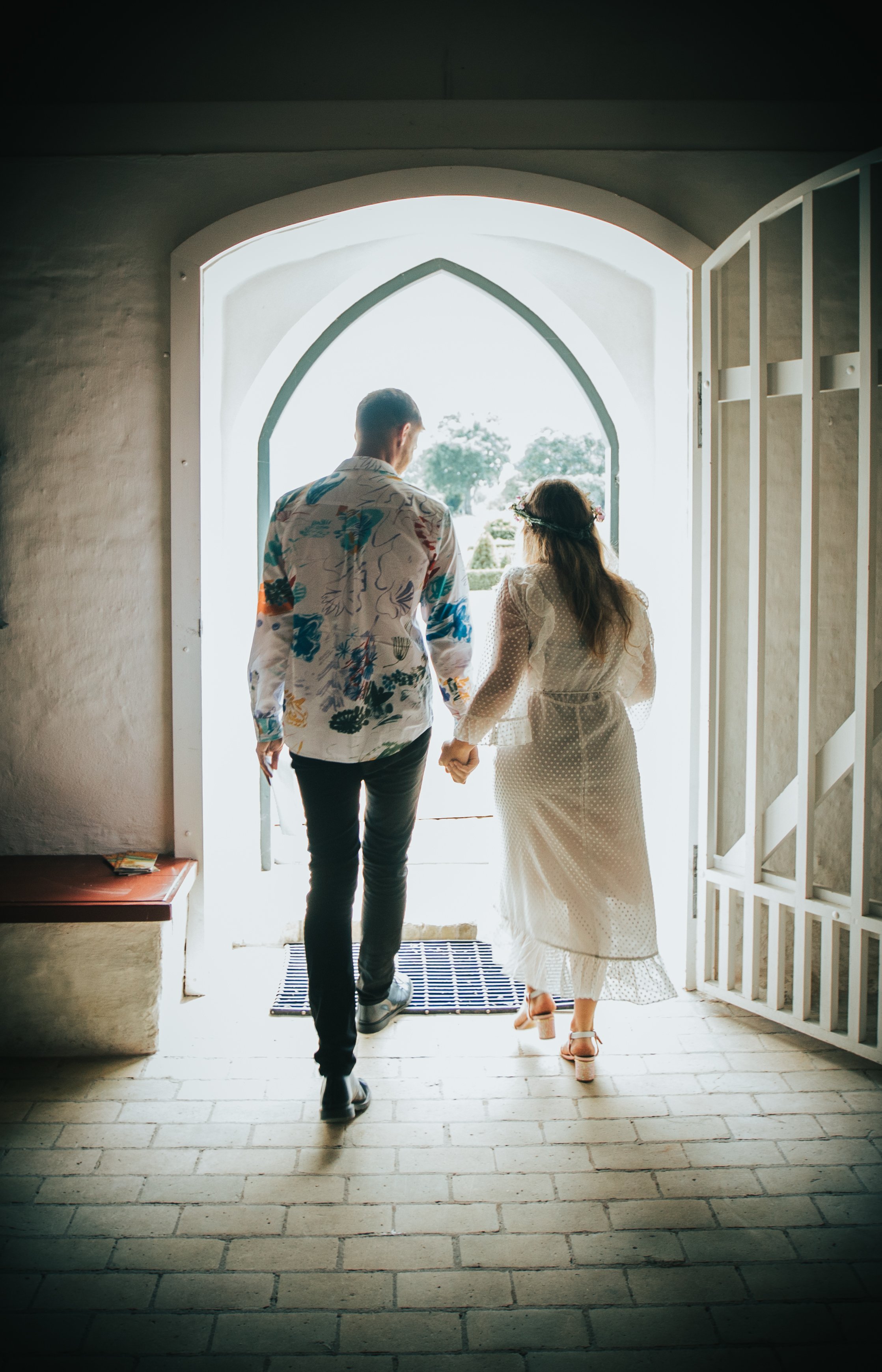 A couple holding hands, walking out of a building through an arched doorway, with bright sunlight and greenery outside.