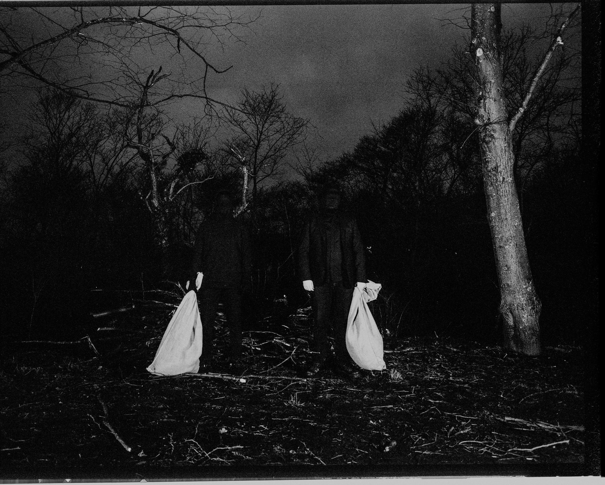 A black and white photo of two people in dark clothing standing in a wooded area at night. Each person is holding a large bag, and there are leafless trees surrounding them under a cloudy sky.