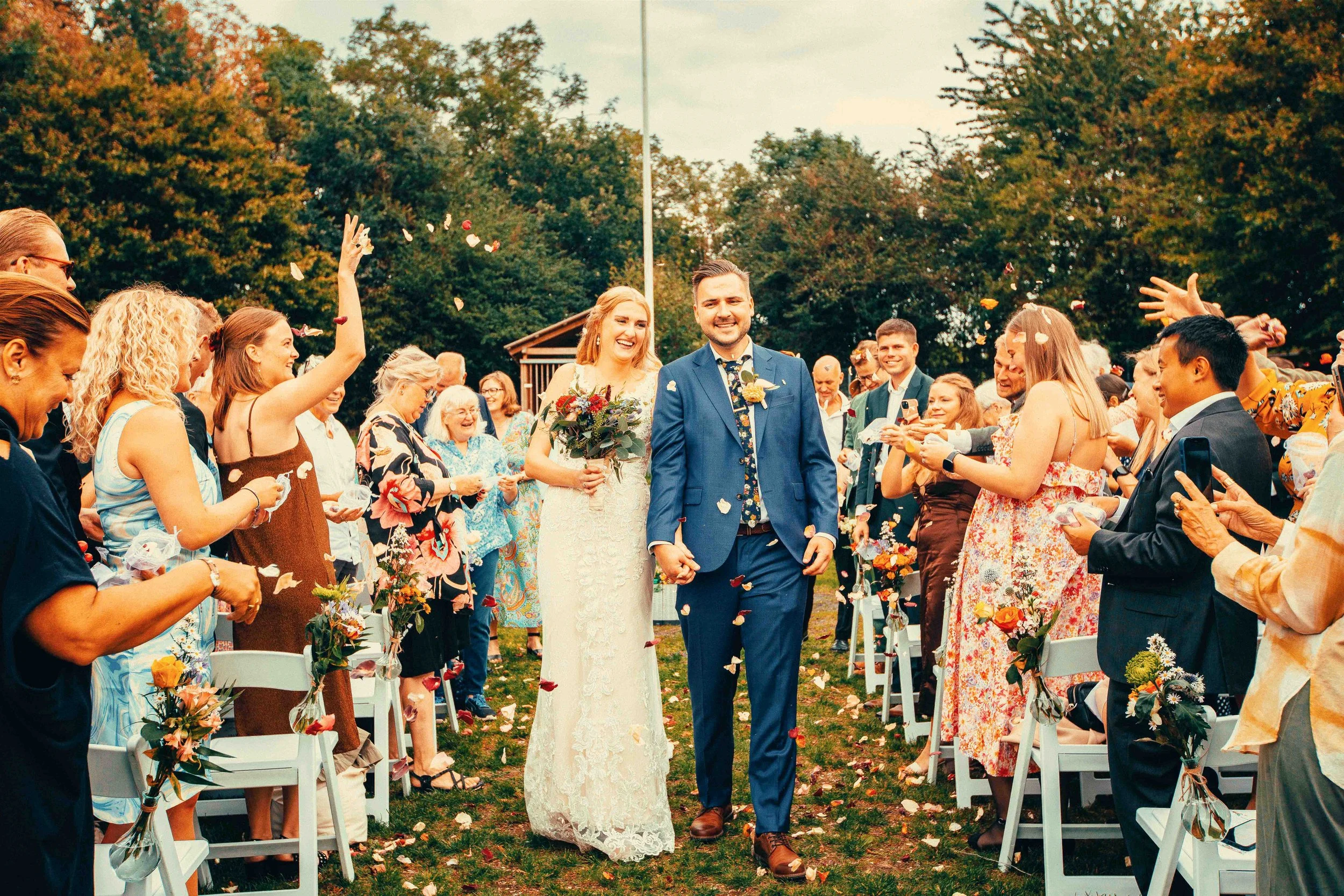 A bride and groom walking together down the aisle at an outdoor wedding ceremony, surrounded by guests throwing flower petals.