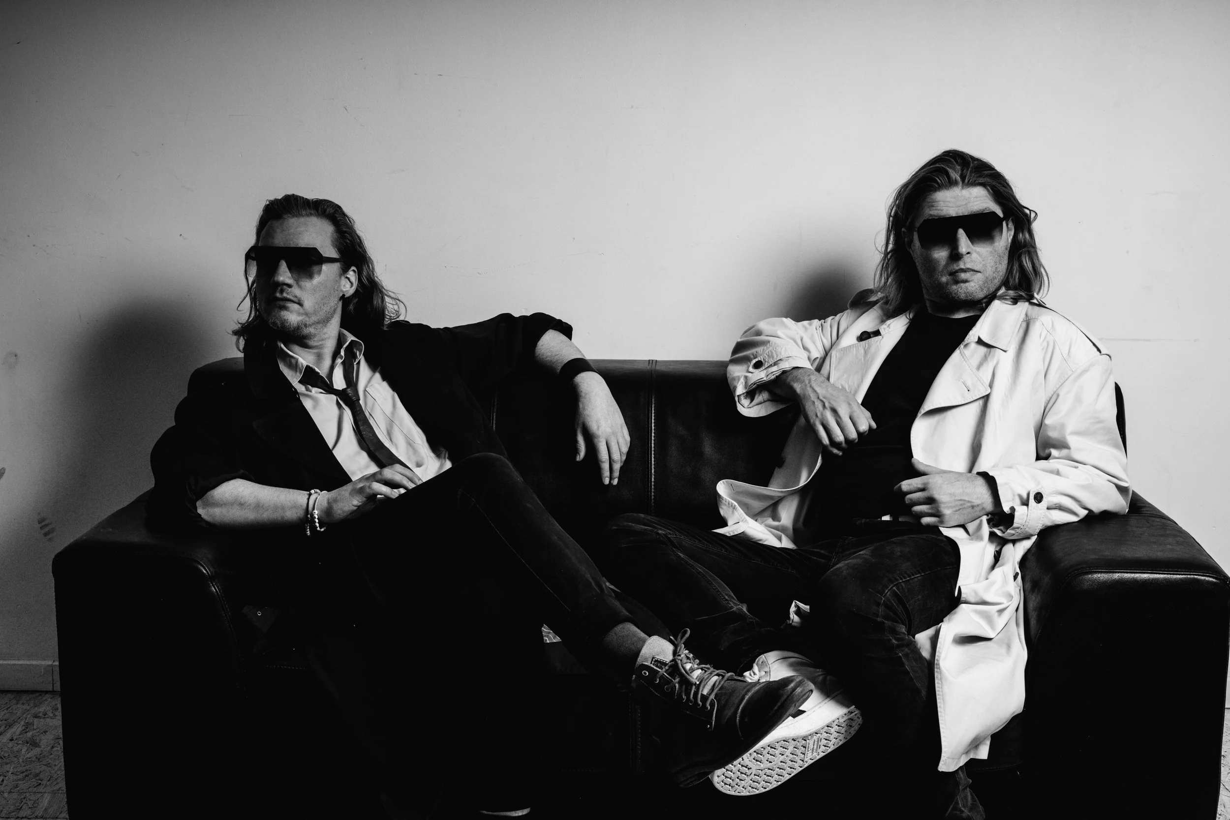 Two men with long hair and sunglasses sitting on a black leather couch against a plain white wall, wearing casual clothing and relaxed poses.