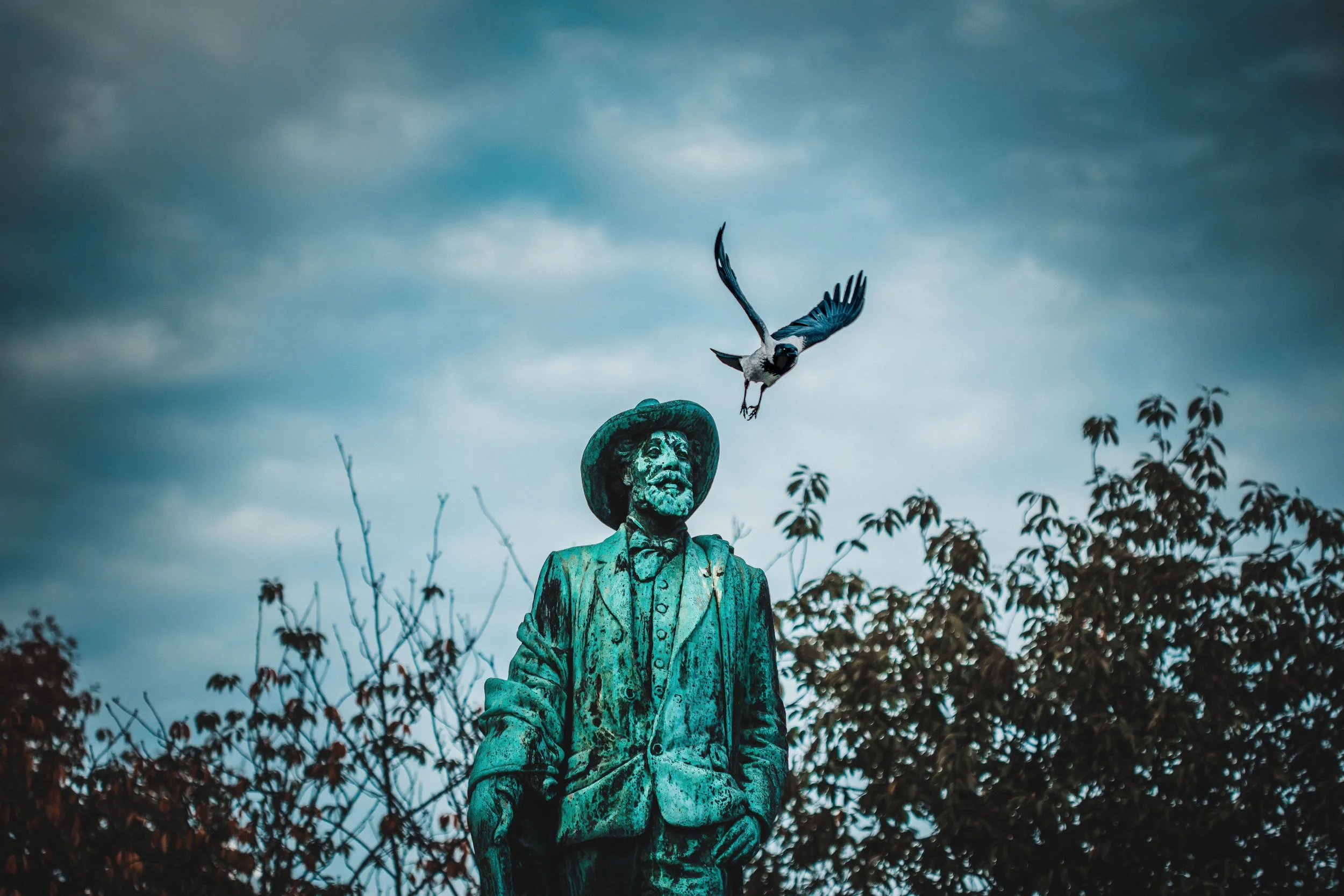 Bronze statue of a man with a beard and hat, surrounded by trees, with a bird flying above against a cloudy sky.